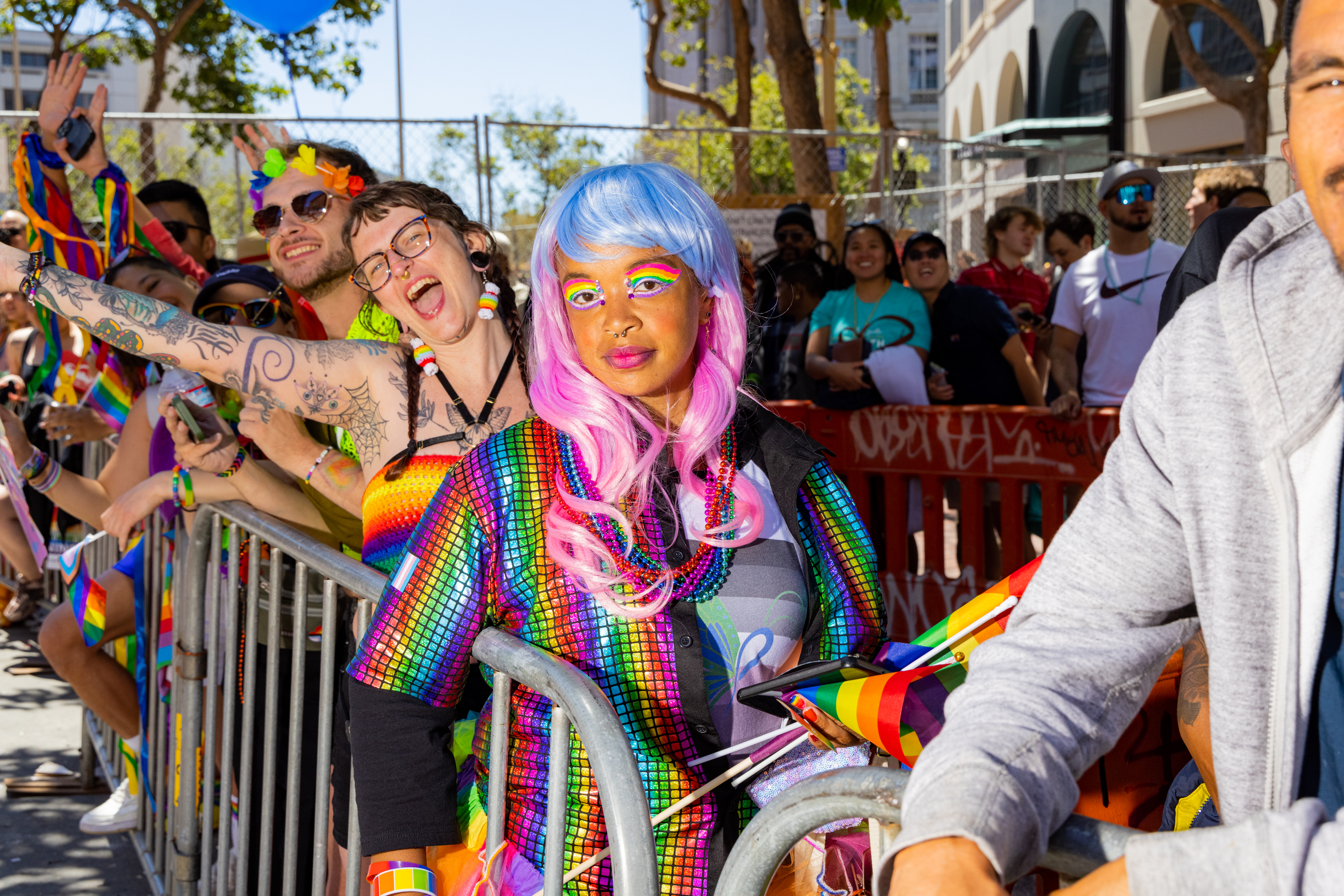 People celebrate at an outdoor event, wearing colorful rainbow attire. A person with a blue and pink wig stands near a barrier, surrounded by cheerful attendees.