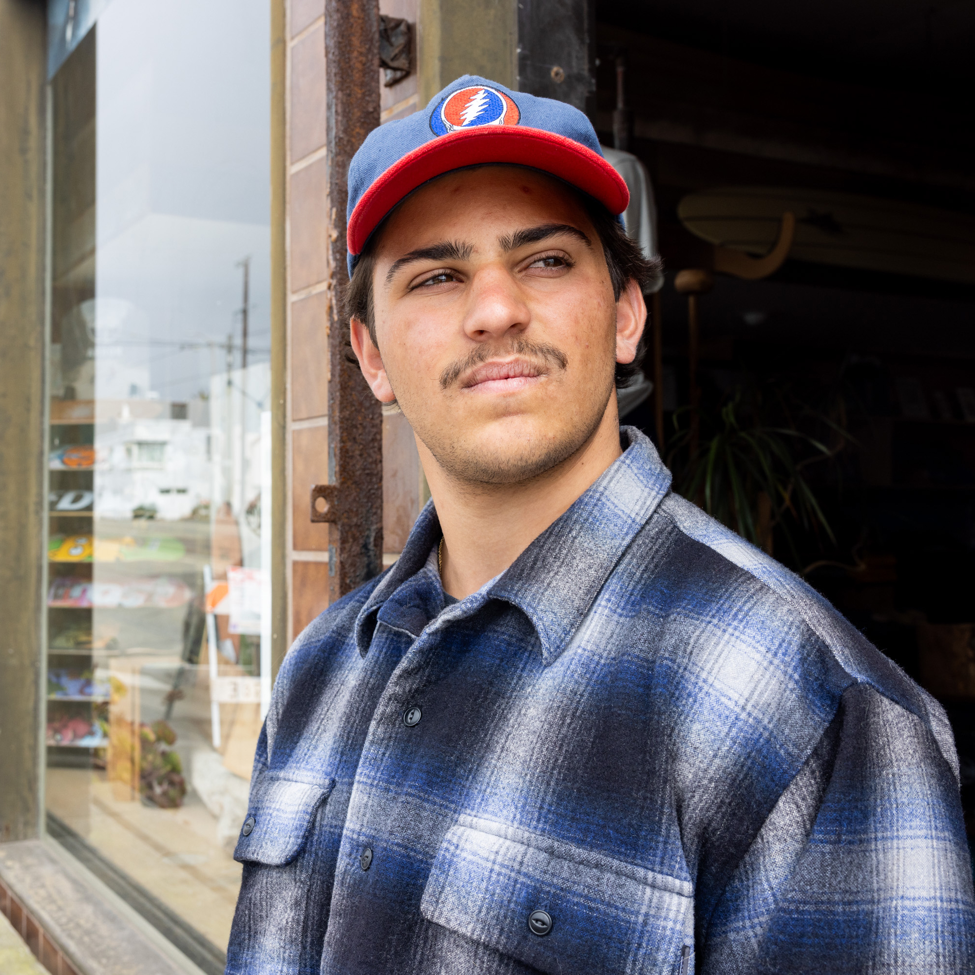 A man with a mustache wears a blue and red cap and a blue plaid shirt, standing outside a shop with a glass window, looking into the distance.