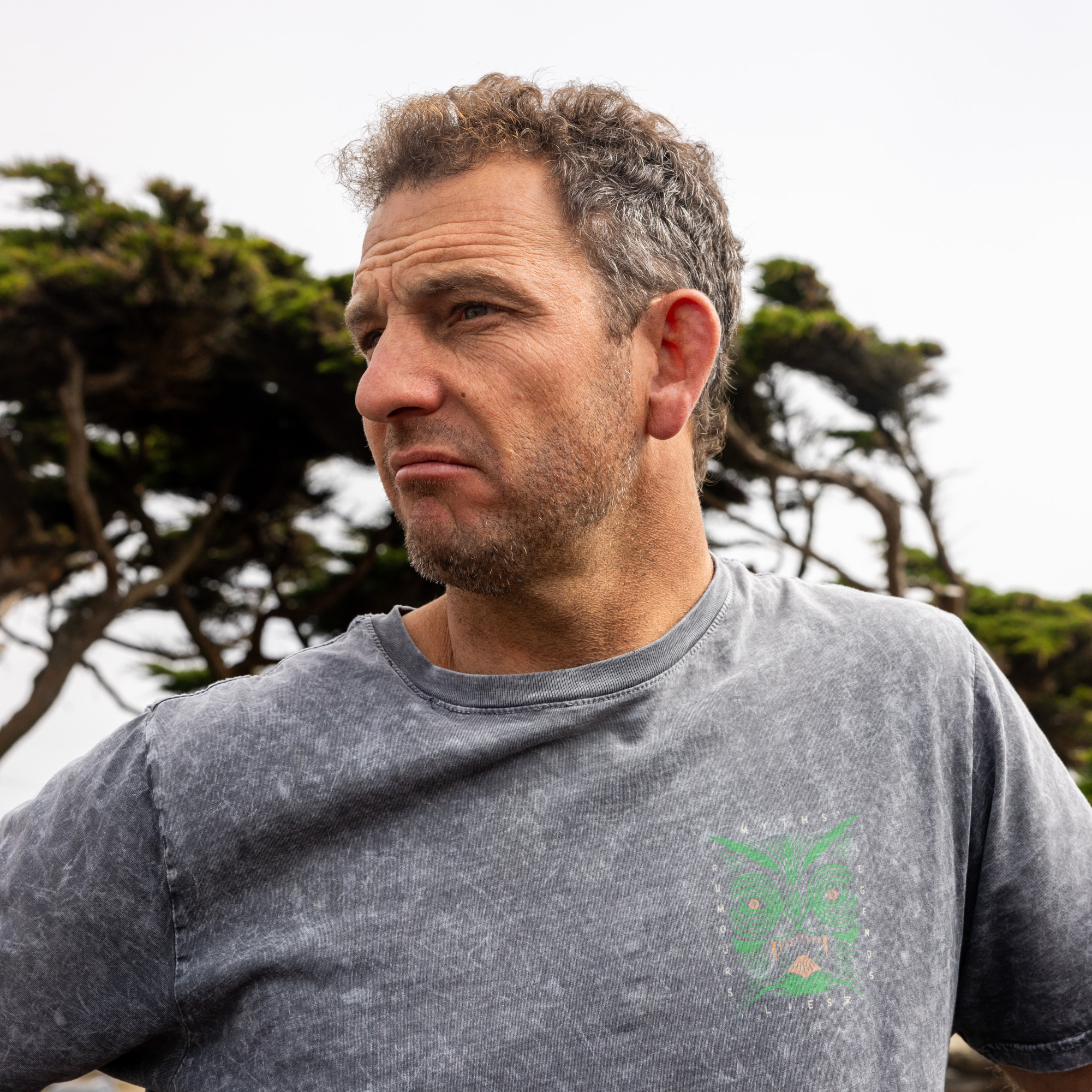 A man with graying curly hair and stubble wears a washed-out gray T-shirt featuring a small green design. He stands outdoors in front of tree branches, looking to the side.