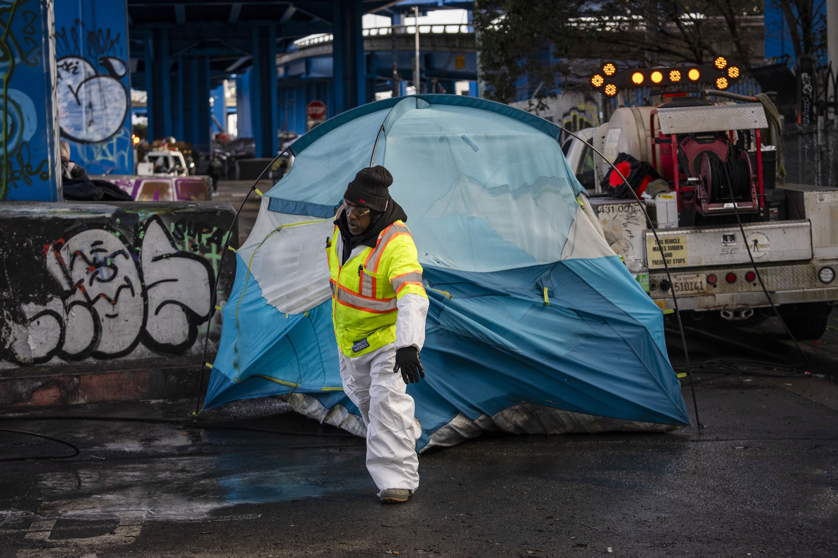 A person in a high-visibility jacket and white protective suit removes a blue tent beneath an overpass with graffiti-covered pillars, and a work truck in the background.