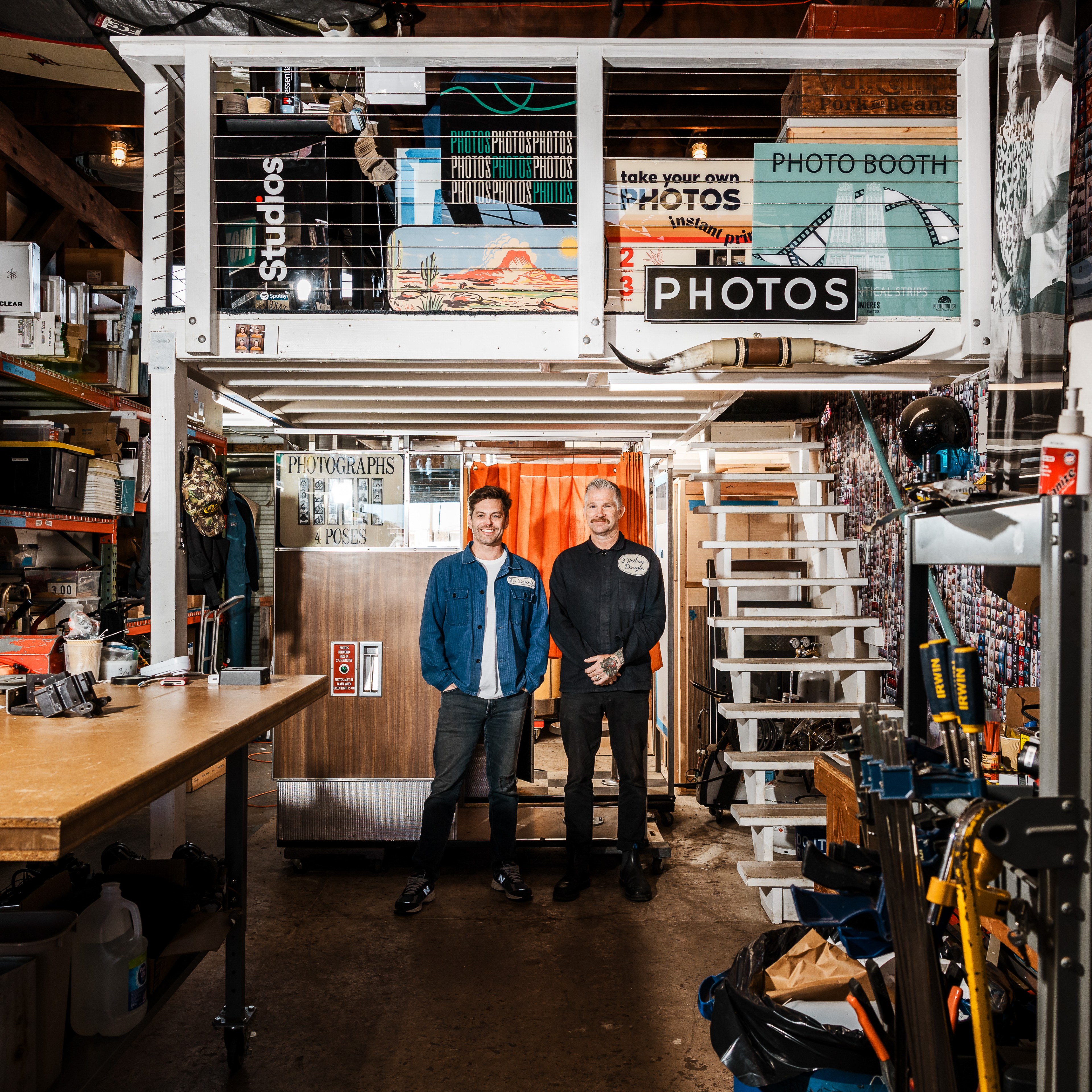 Two men stand in a cluttered workshop with a photo booth, surrounded by tools, equipment, and colorful signs advertising photos on a mezzanine above.