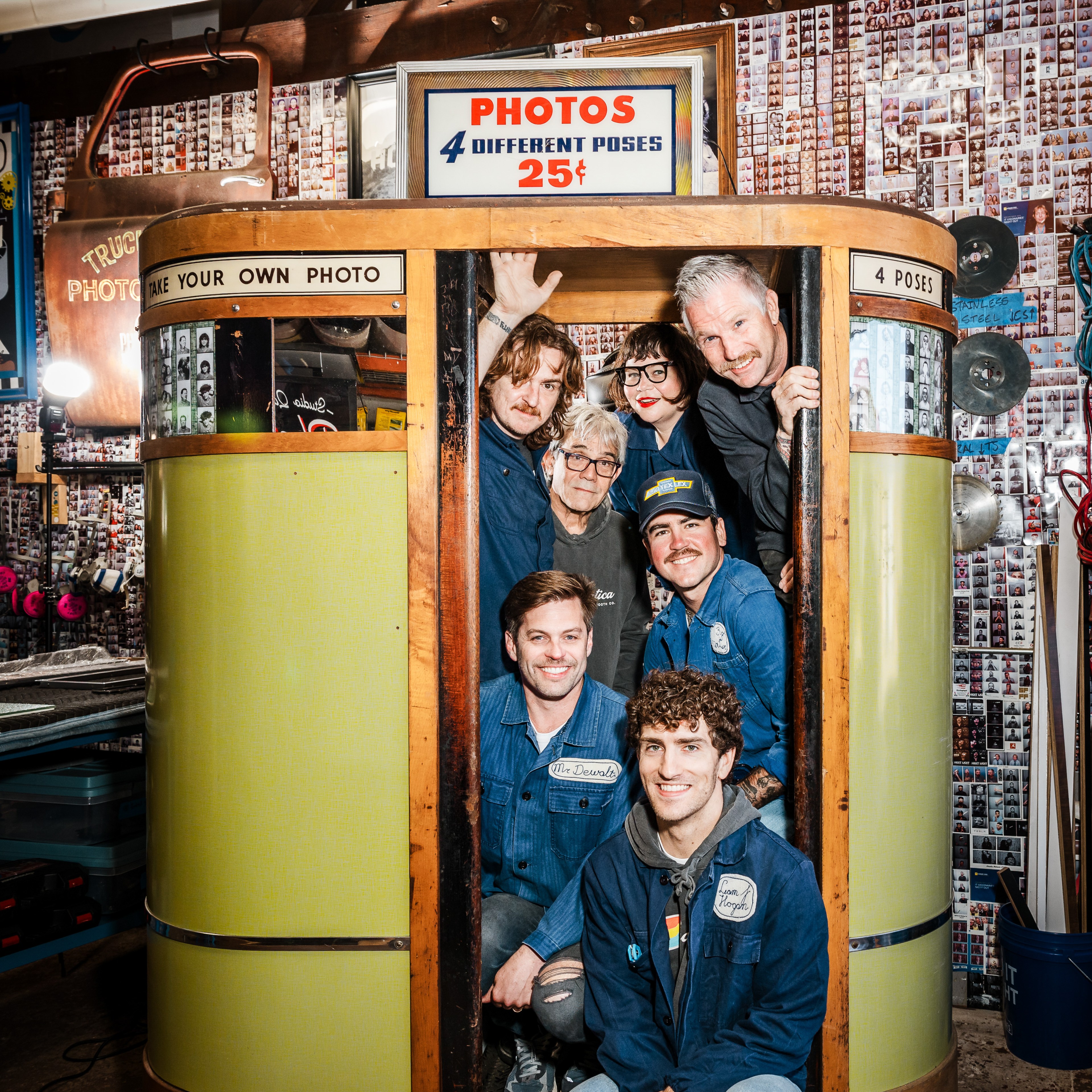 Seven people are crowded in an old-fashioned photo booth, smiling. The booth advertises “PHOTOS 4 DIFFERENT POSES 25¢.” Walls are filled with photo strips.