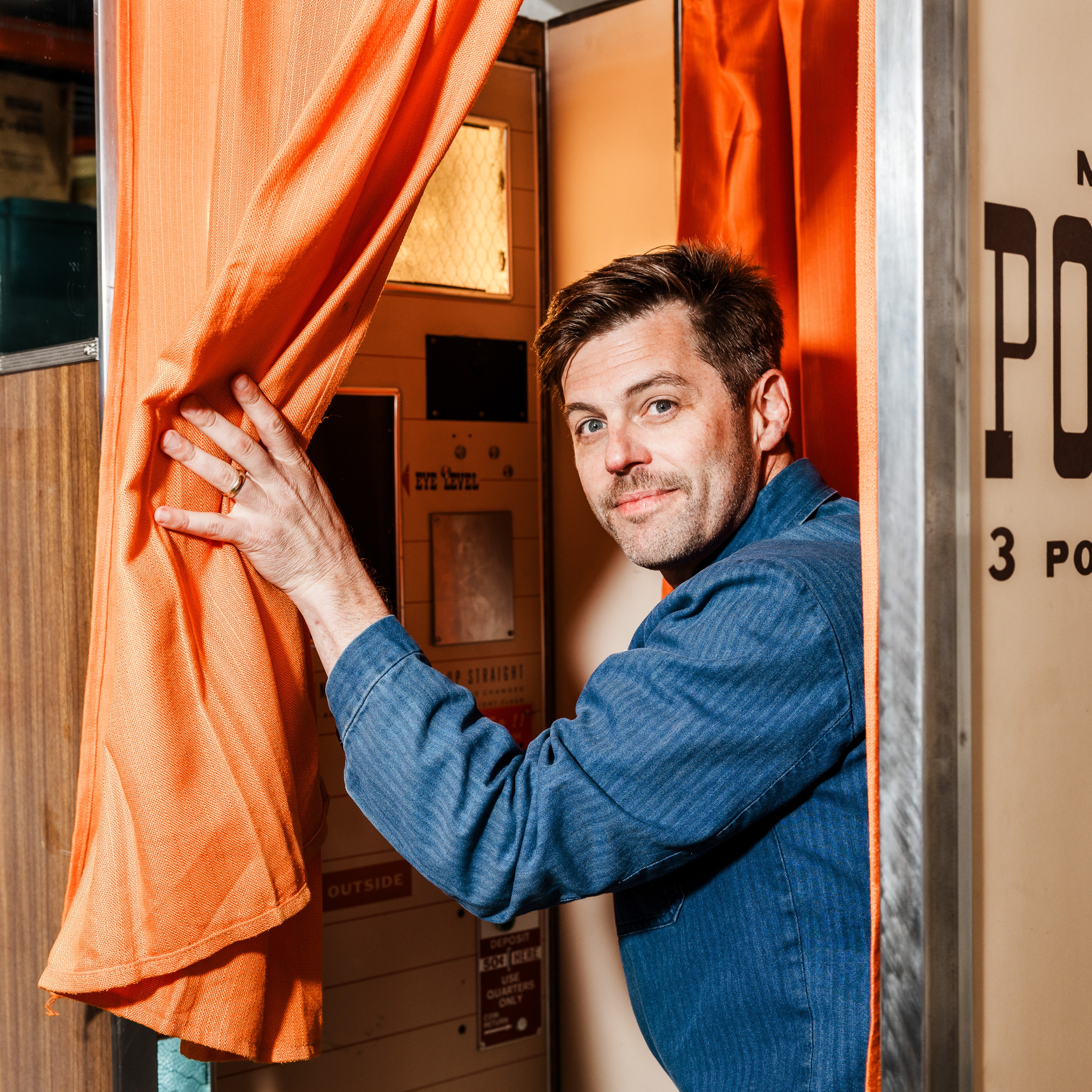 A man with short, dark hair and a light stubble peeks out from behind an orange curtain, holding it open with one hand. He stands inside a photo booth.