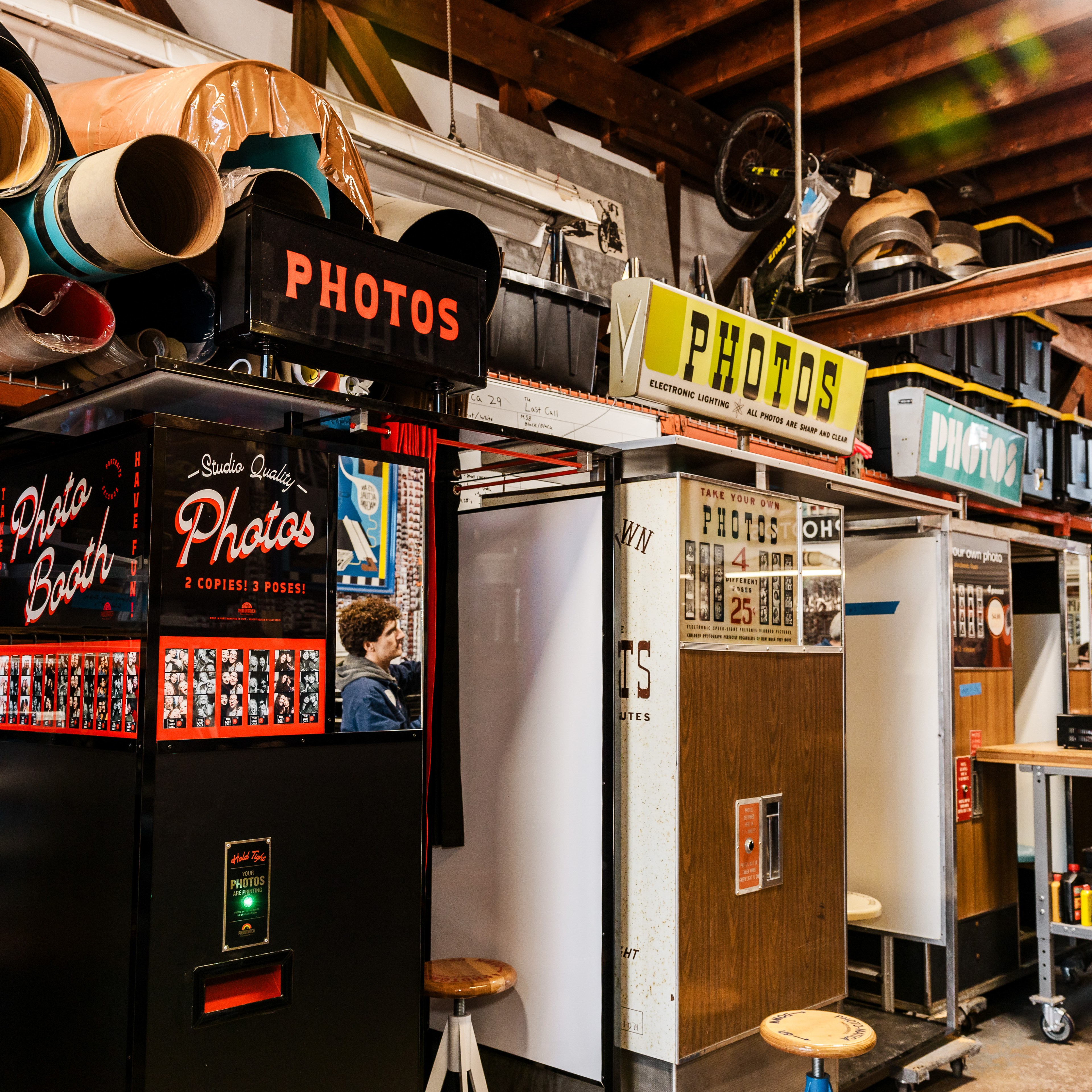 The image shows a room filled with vintage photo booths, each bearing signs that say &quot;PHOTOS&quot;. The room is cluttered with photo rolls and has a wooden ceiling.