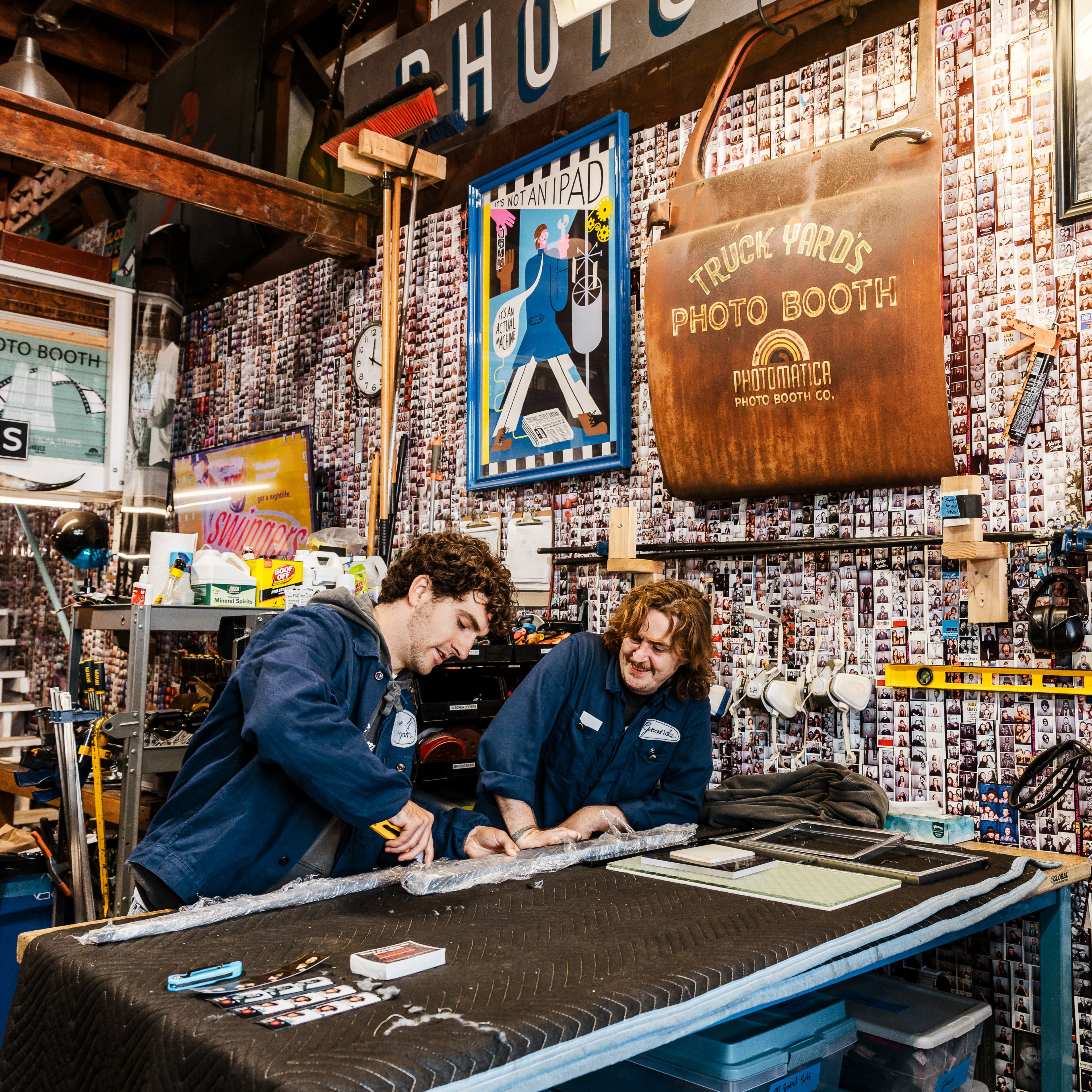 Two people in blue jackets work together in a cluttered workshop. The walls are covered with photo strips, various tools, and colorful posters.
