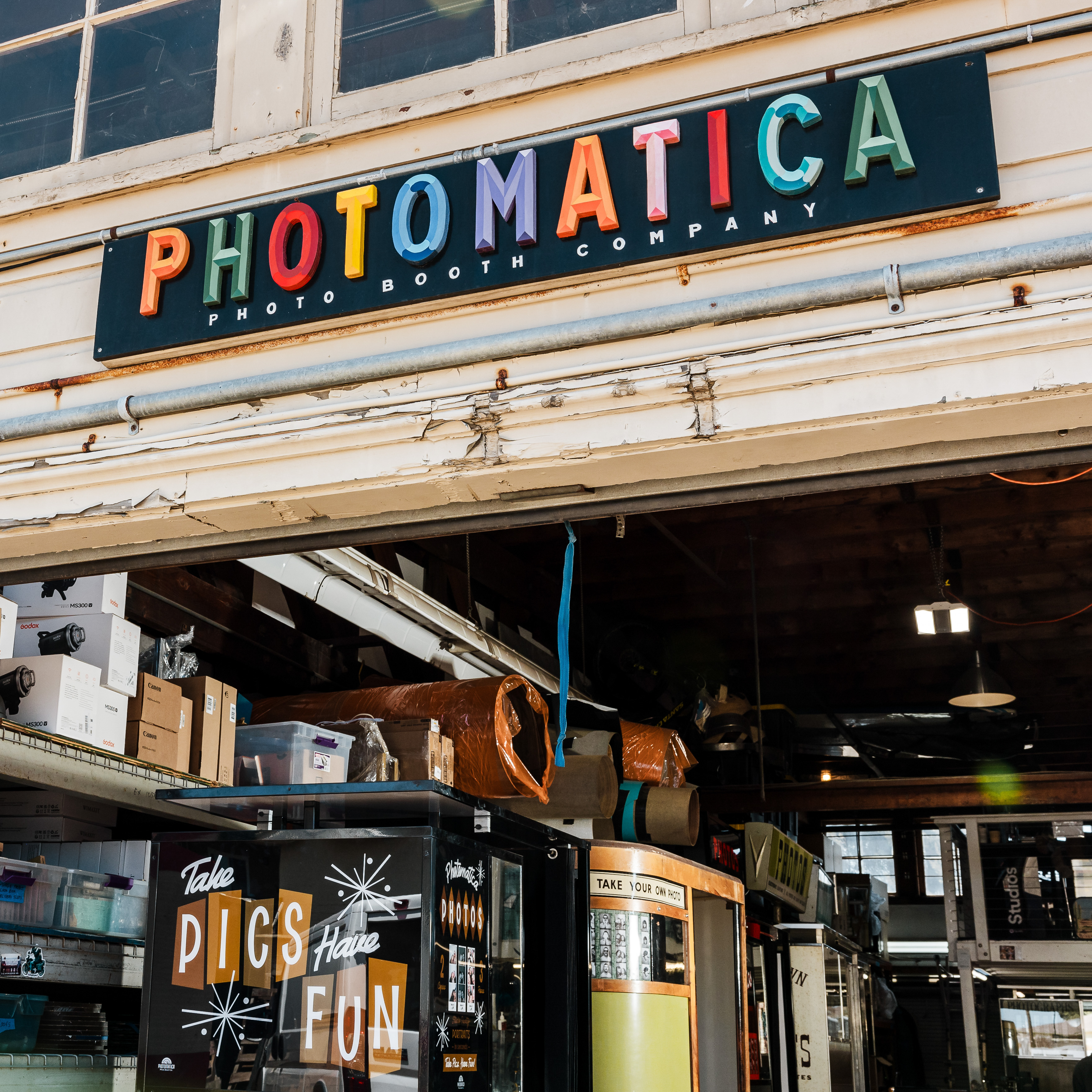 The image shows a store front with a sign that reads &quot;PHOTOMATICA&quot; in colorful letters. Below, there is a photo booth with the words &quot;Take Pics Have Fun.&quot; Various items are stored above.