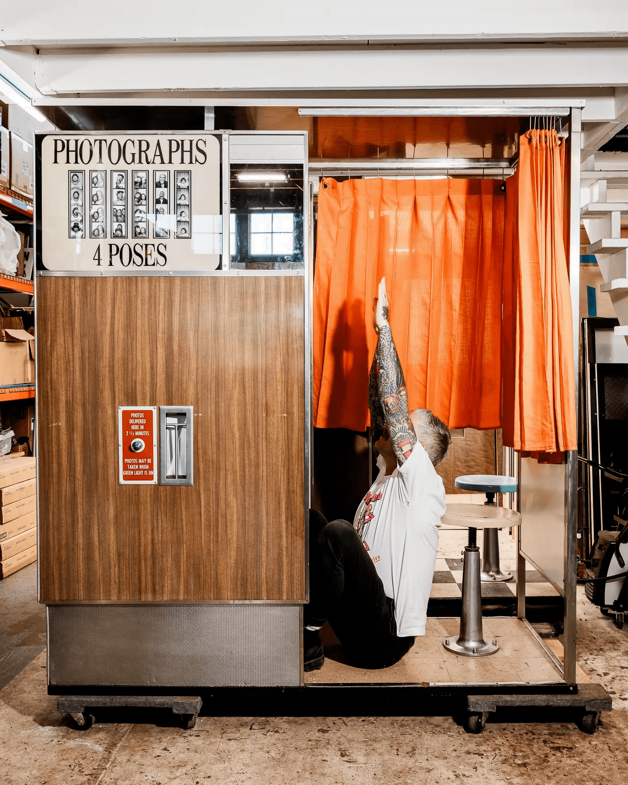 A person with tattooed arms, wearing a white shirt, is sitting and stretching inside a vintage photo booth with an orange curtain, in an industrial setting.
