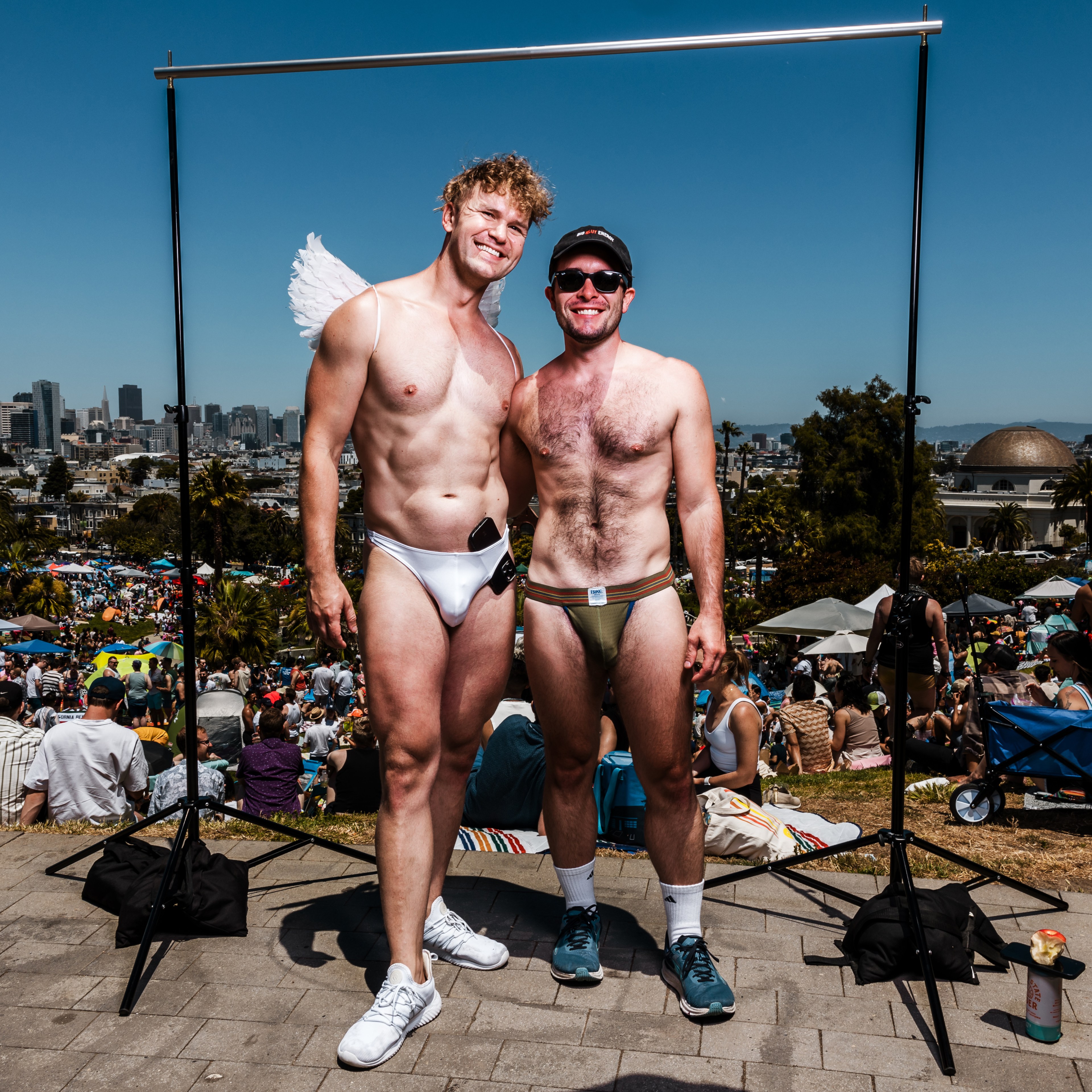 Two men in minimal swimwear stand smiling in front of a crowded outdoor park, framed by a metal stand, with a city skyline and blue sky in the background.