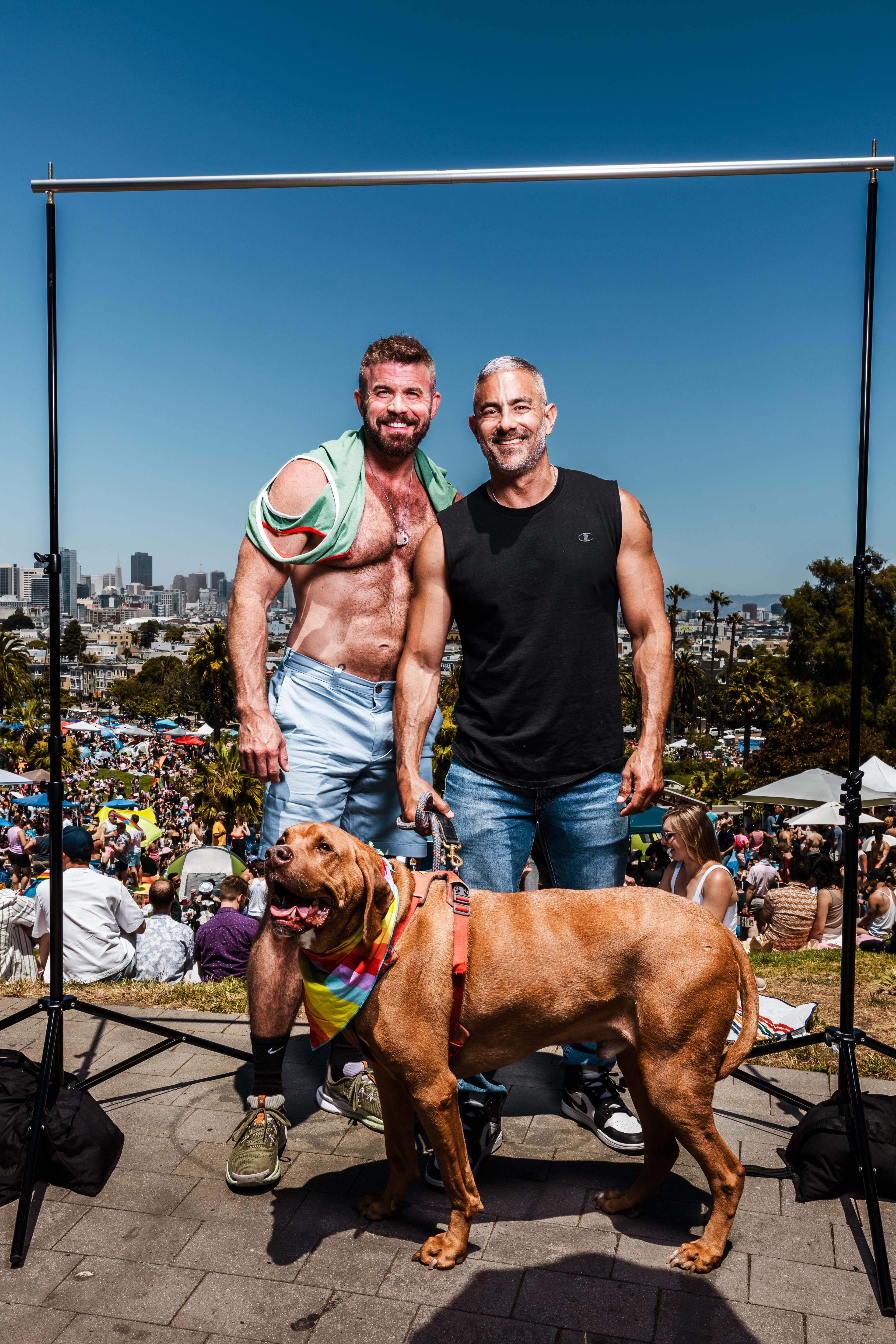 Two men stand smiling with a large dog wearing a rainbow bandana in front of a crowded park with city buildings in the background under a clear blue sky.