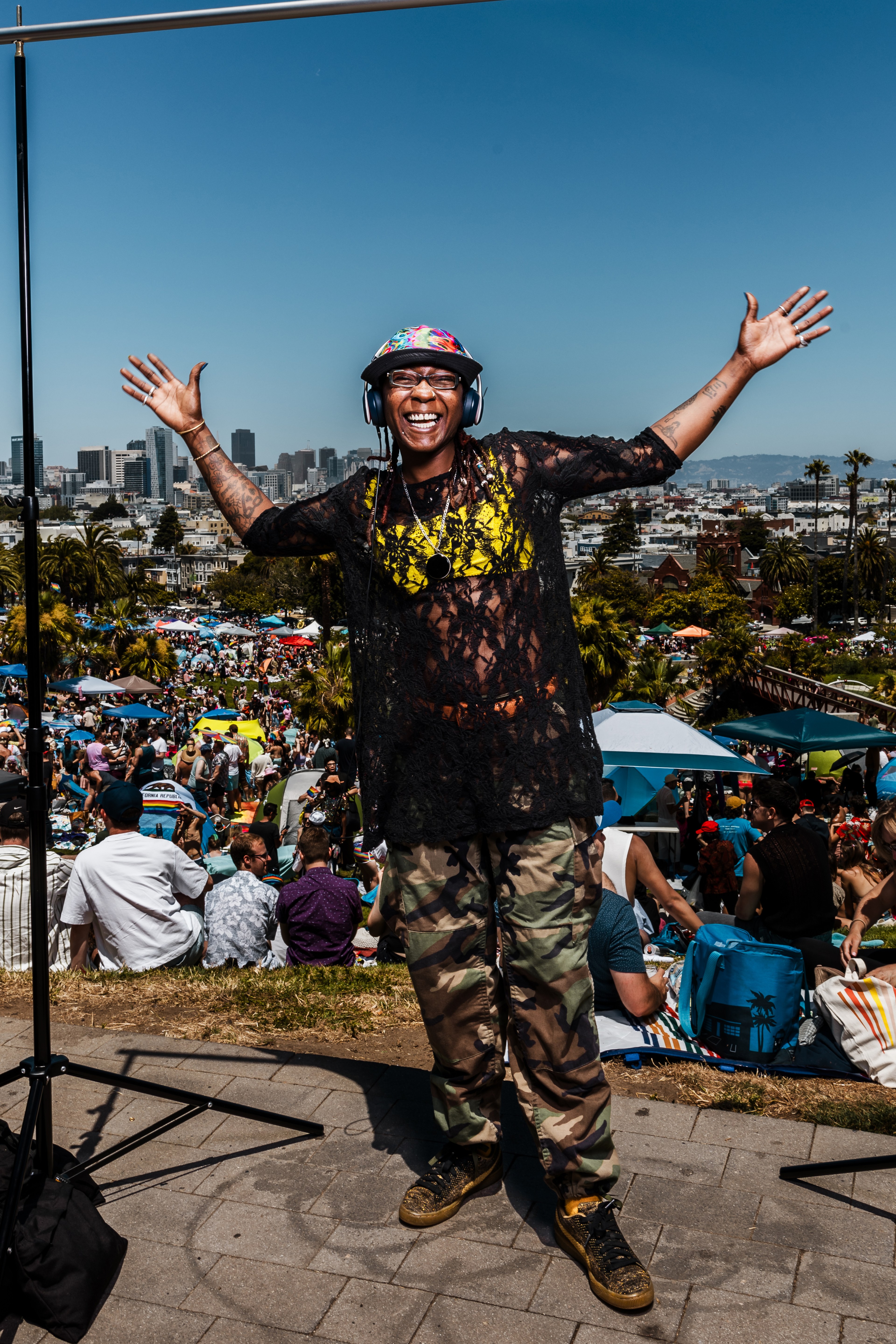 A person with a colorful cap and headphones, wearing a black netted top and camouflage pants, spreads their arms wide, smiling in front of a large festive crowd and cityscape.