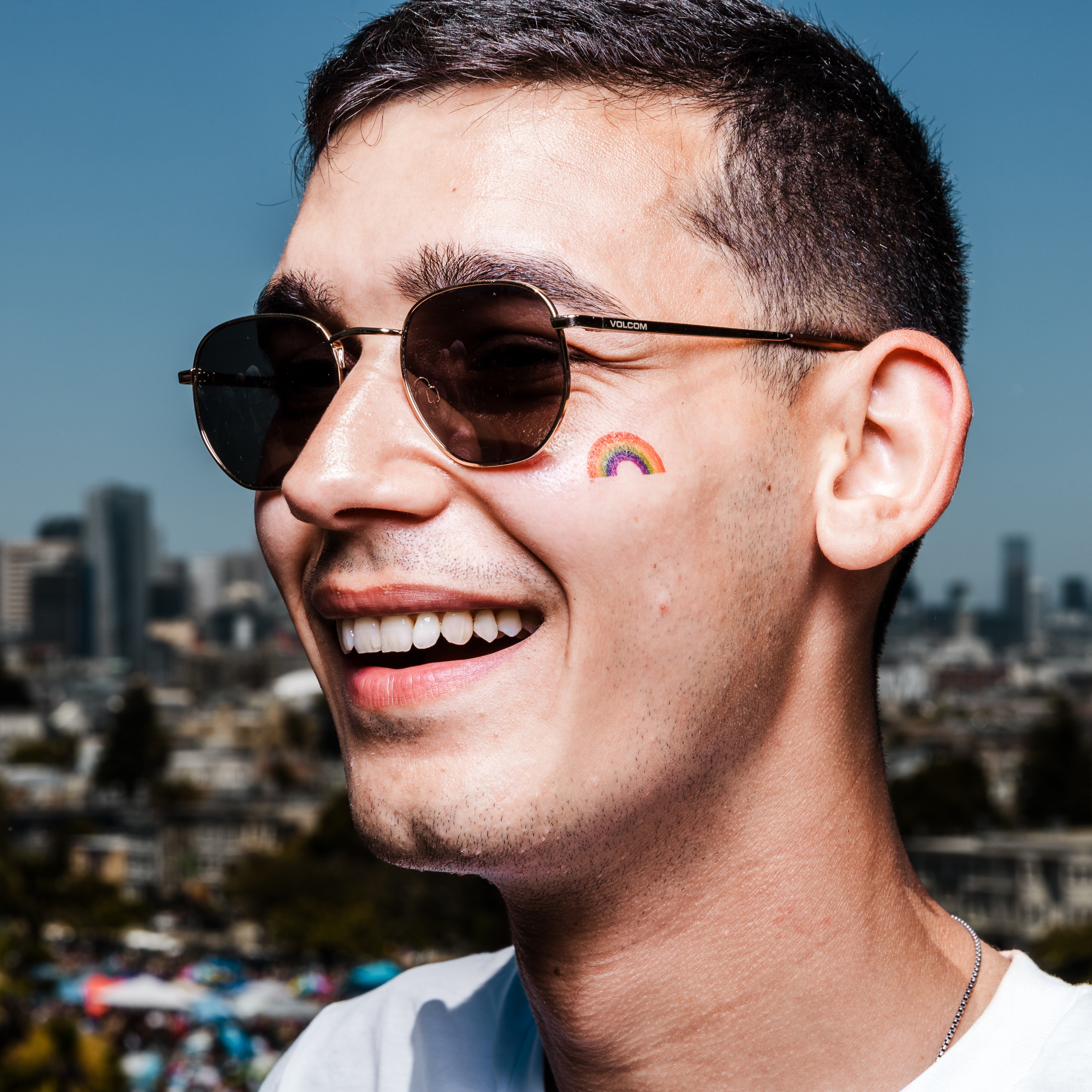 A smiling person with short hair and sunglasses has a small rainbow sticker on their cheek, with a cityscape backdrop under a clear blue sky.