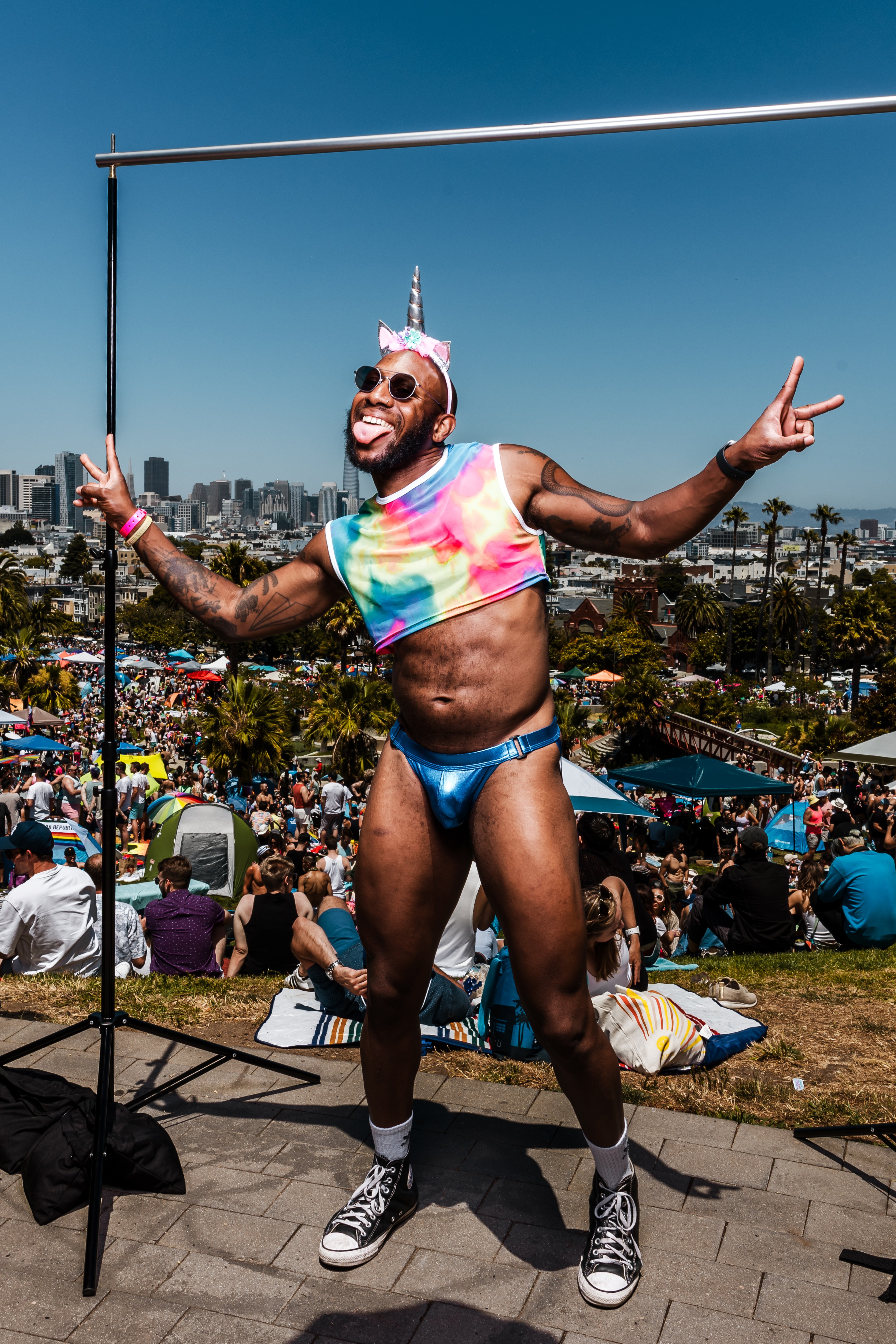 A person dressed in a rainbow crop top, blue briefs, and a unicorn headband with a horn poses joyfully, making a peace sign. They are in a park crowded with people.