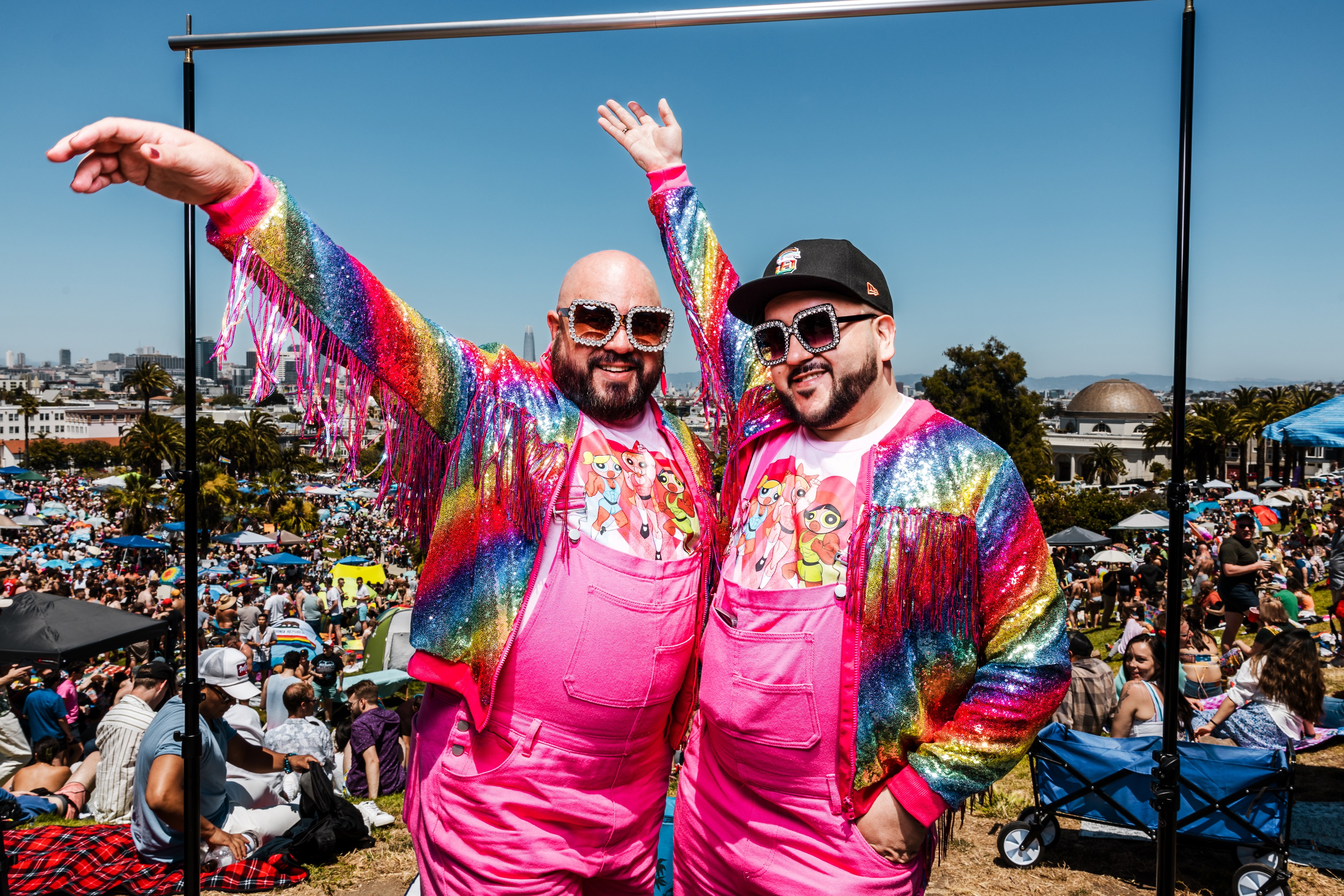 Two smiling men in matching colorful, sequined jackets and pink overalls pose joyfully with arms raised. They wear large sunglasses. A lively outdoor crowd is in the background.