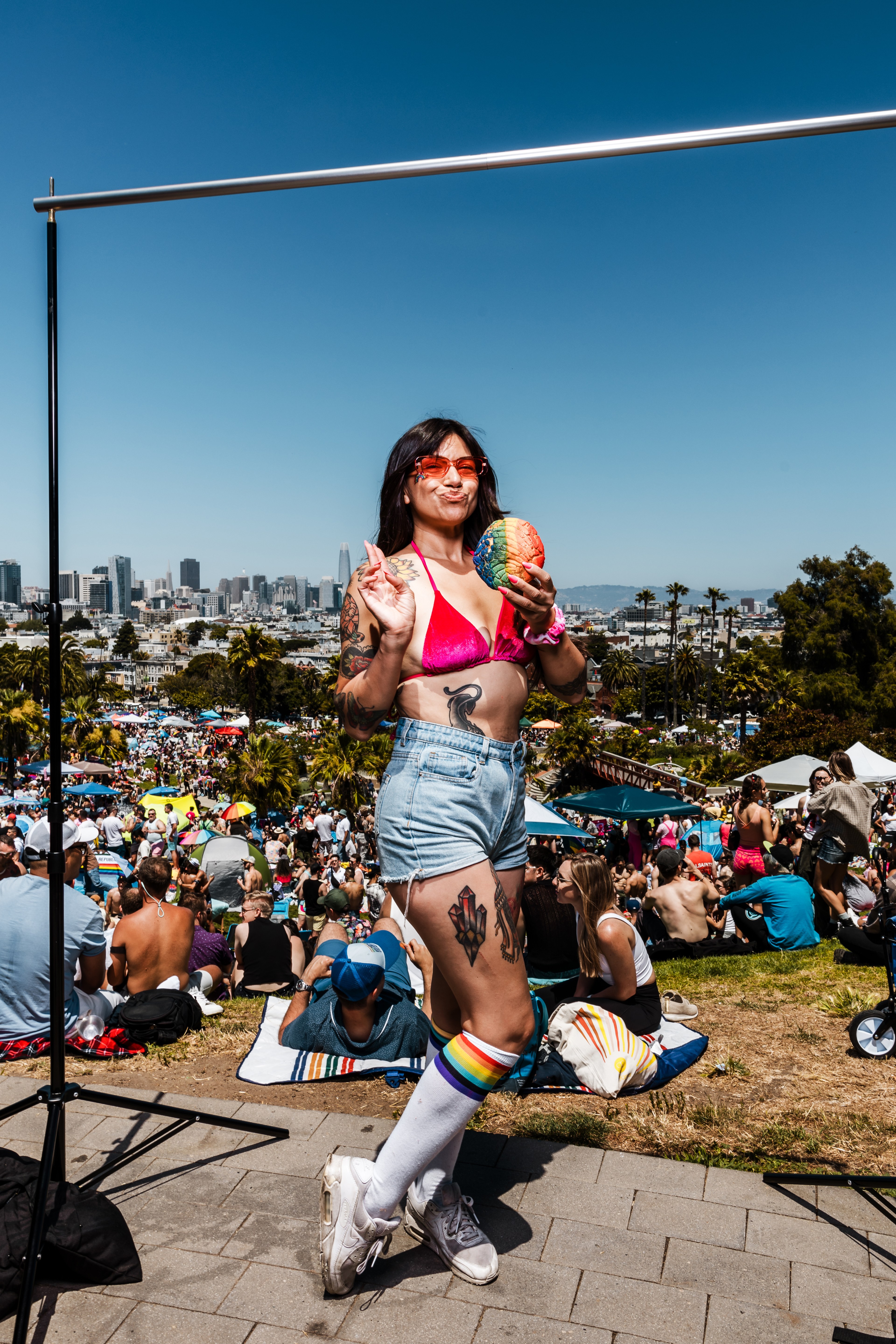 A woman in a pink bikini top, denim shorts, and rainbow socks poses with a peace sign and a colorful object, surrounded by a festive crowd in a sunny outdoor setting.