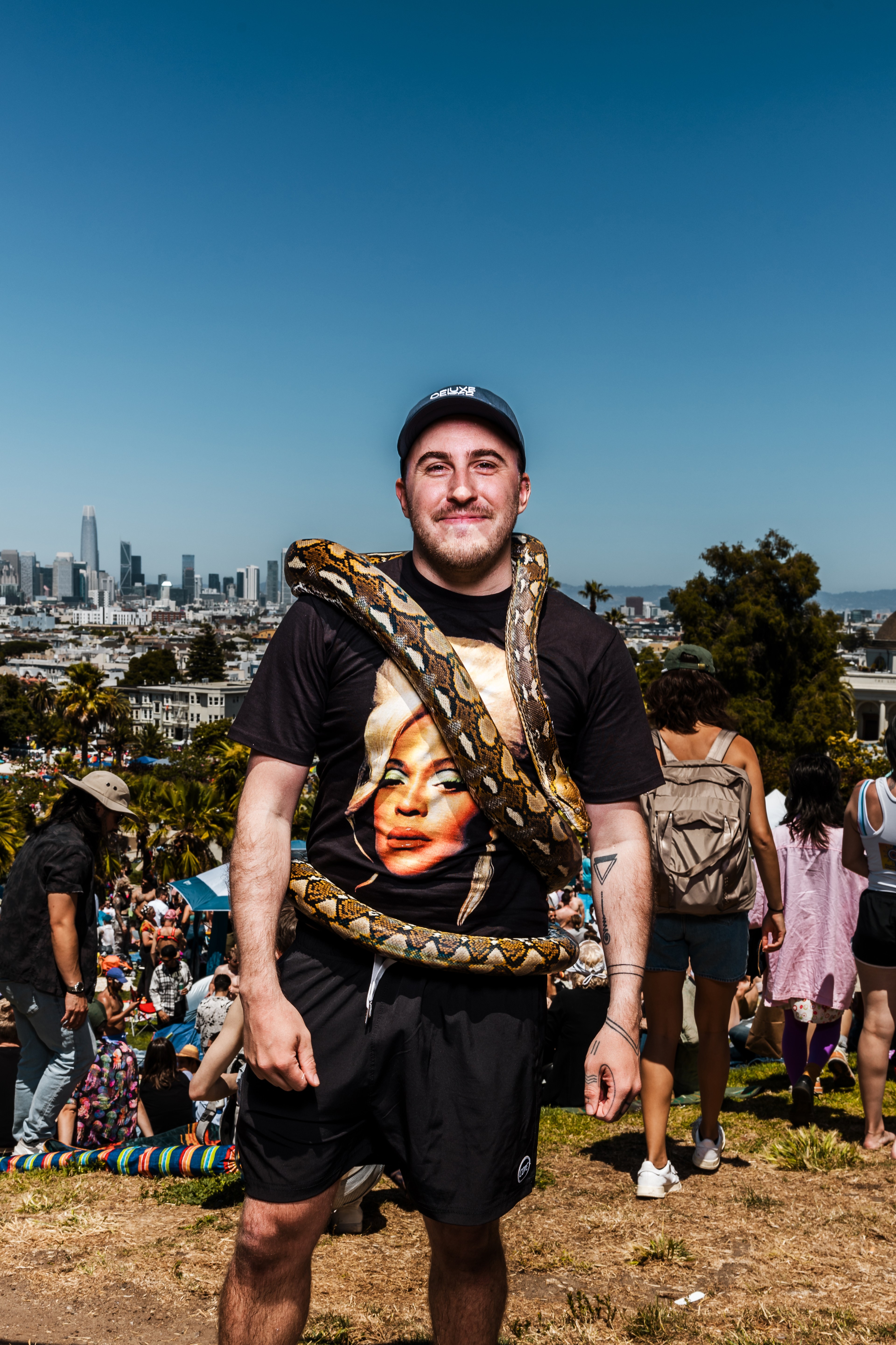 A man stands outdoors with a large snake draped over his shoulders. He wears a hat and a printed black t-shirt. People are gathered in a park under a clear sky.