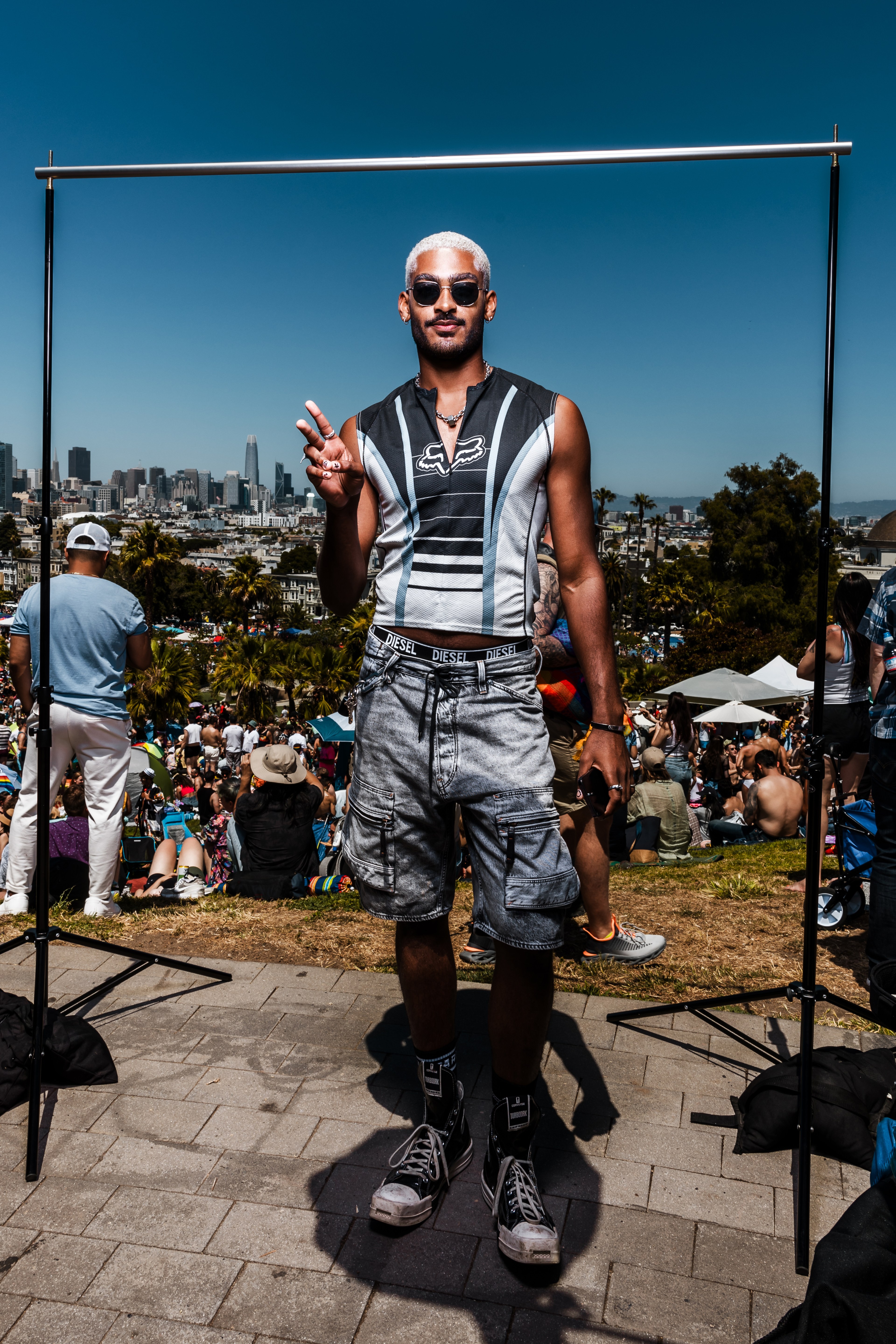 A person stands outdoors in front of a crowd, making a peace sign. They wear sunglasses, a sleeveless top, cargo shorts, and high-top sneakers, with a city skyline behind them.