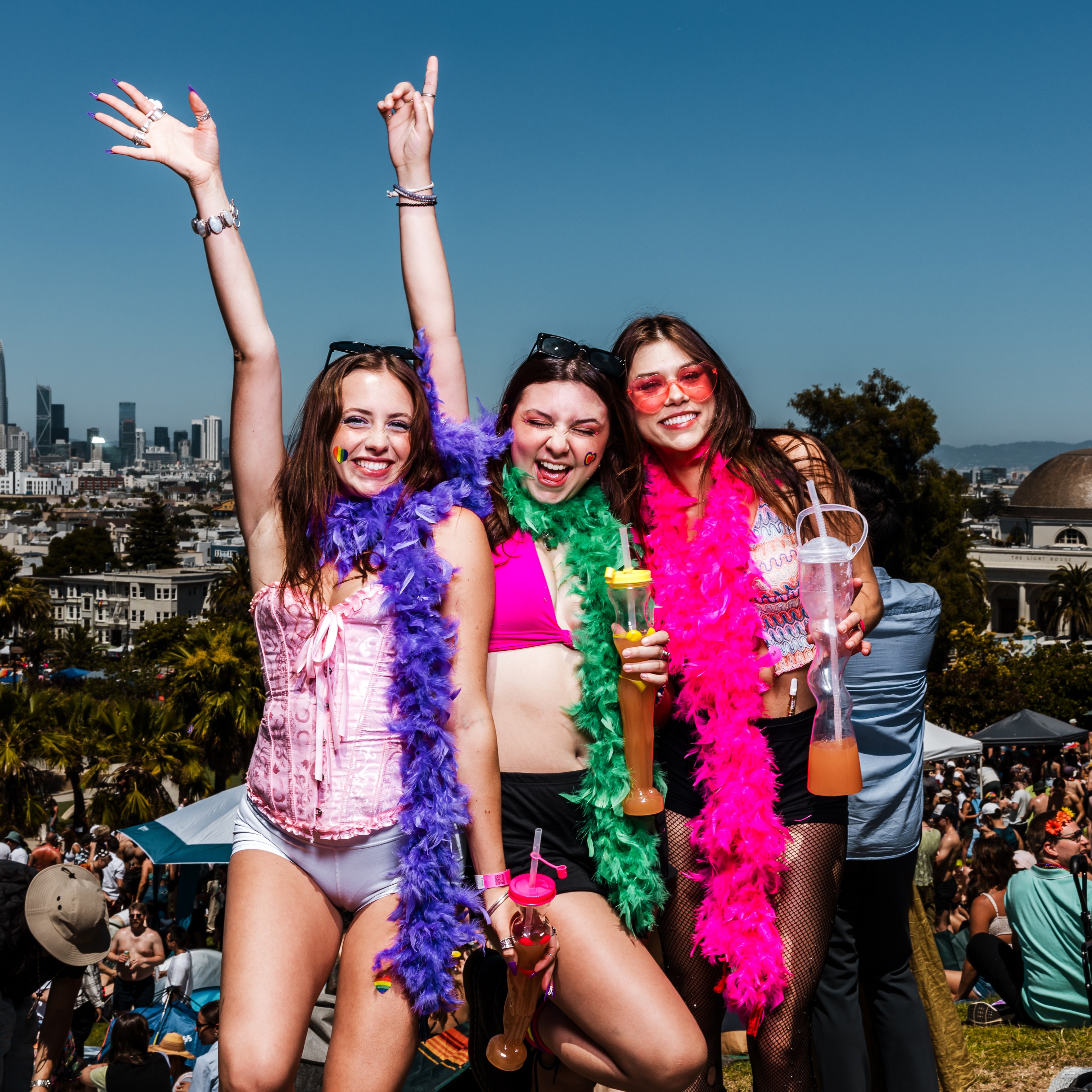 Three women wearing colorful feather boas and festival outfits pose cheerfully with drinks in hand at an outdoor event, with a cityscape in the background.