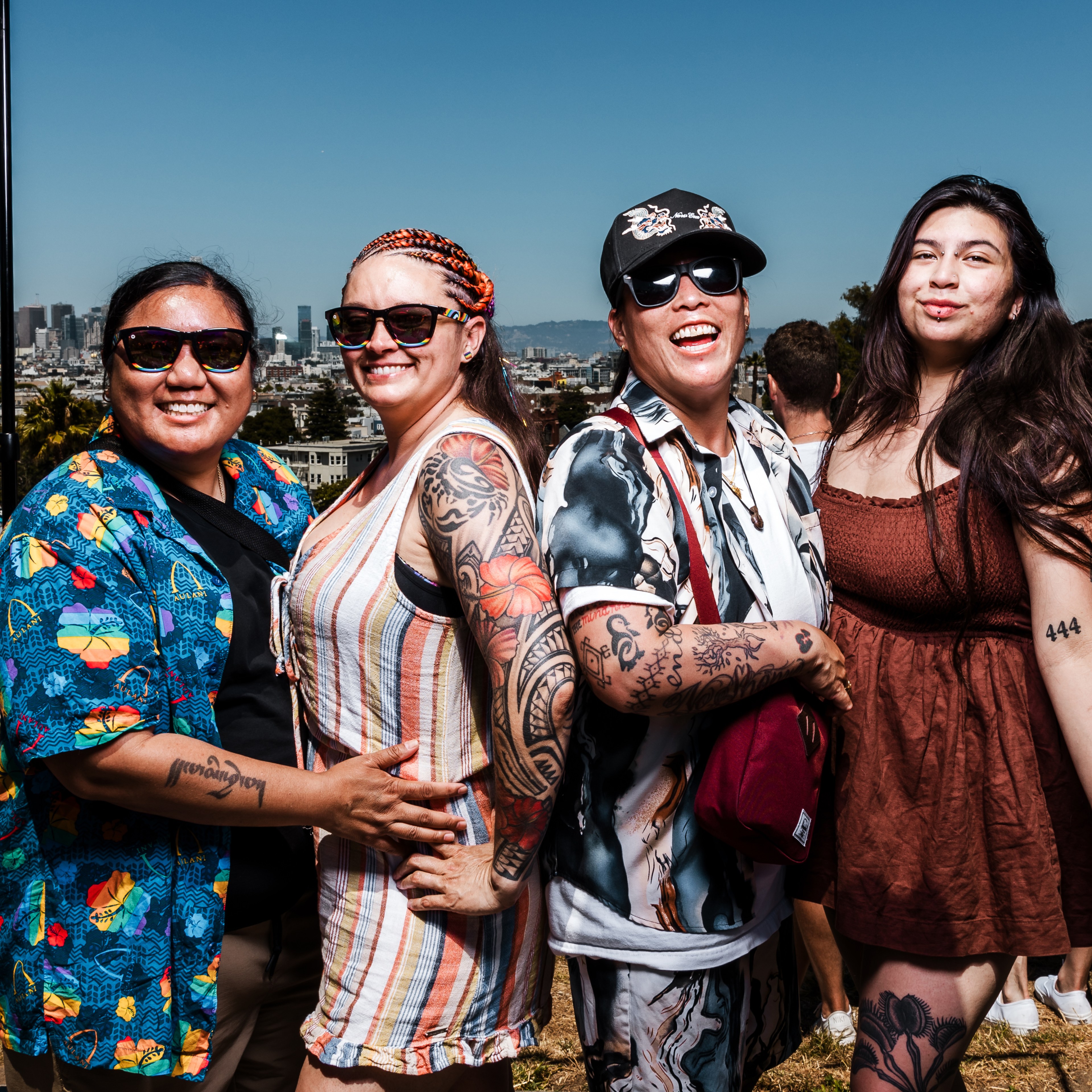 Four friends, all wearing sunglasses, stand close together smiling. They have diverse outfits and visible tattoos. A cityscape is in the background under a clear blue sky.