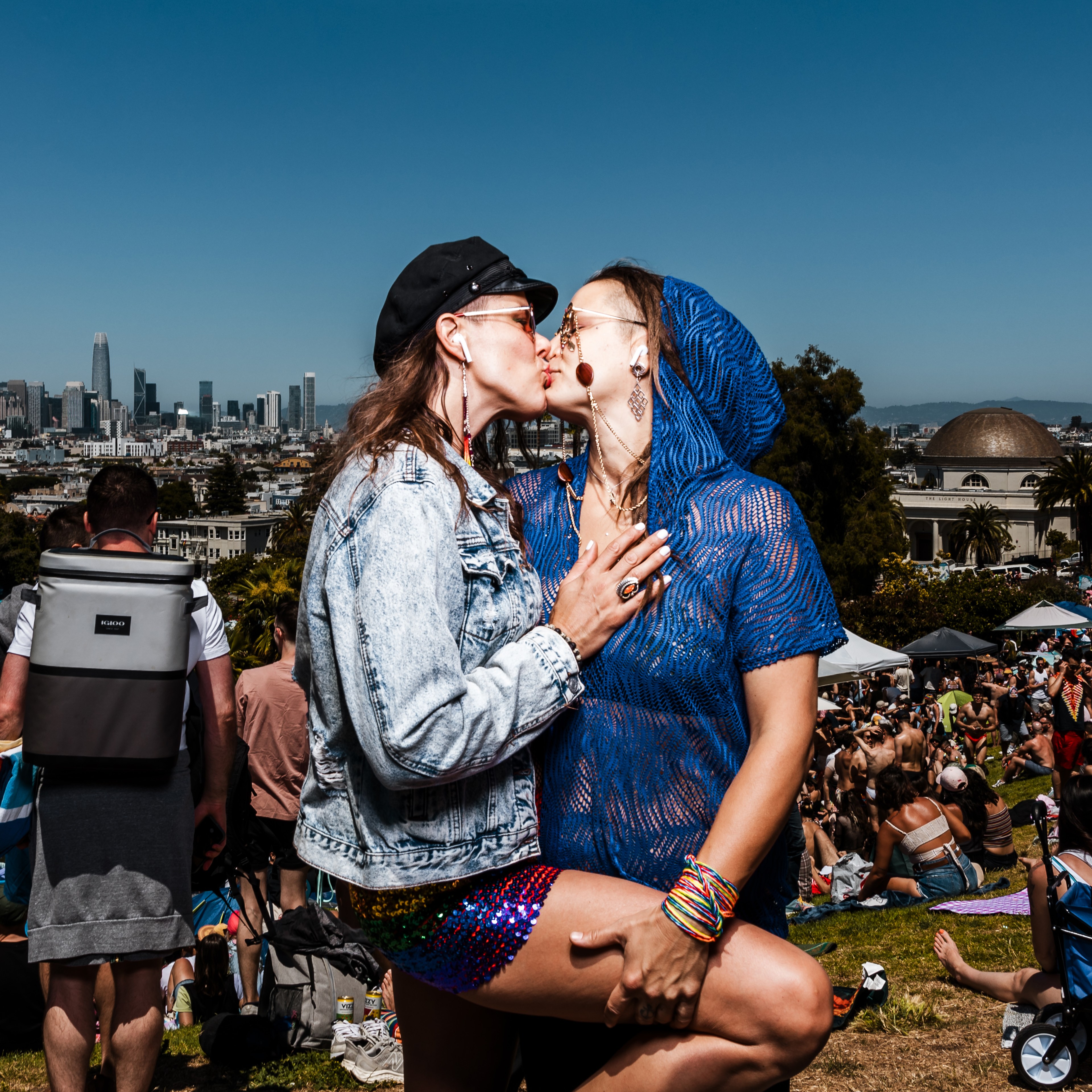 Two people share a kiss at an outdoor event with a crowd behind them and a city skyline in the background. One wears a denim jacket, and the other a blue mesh top.