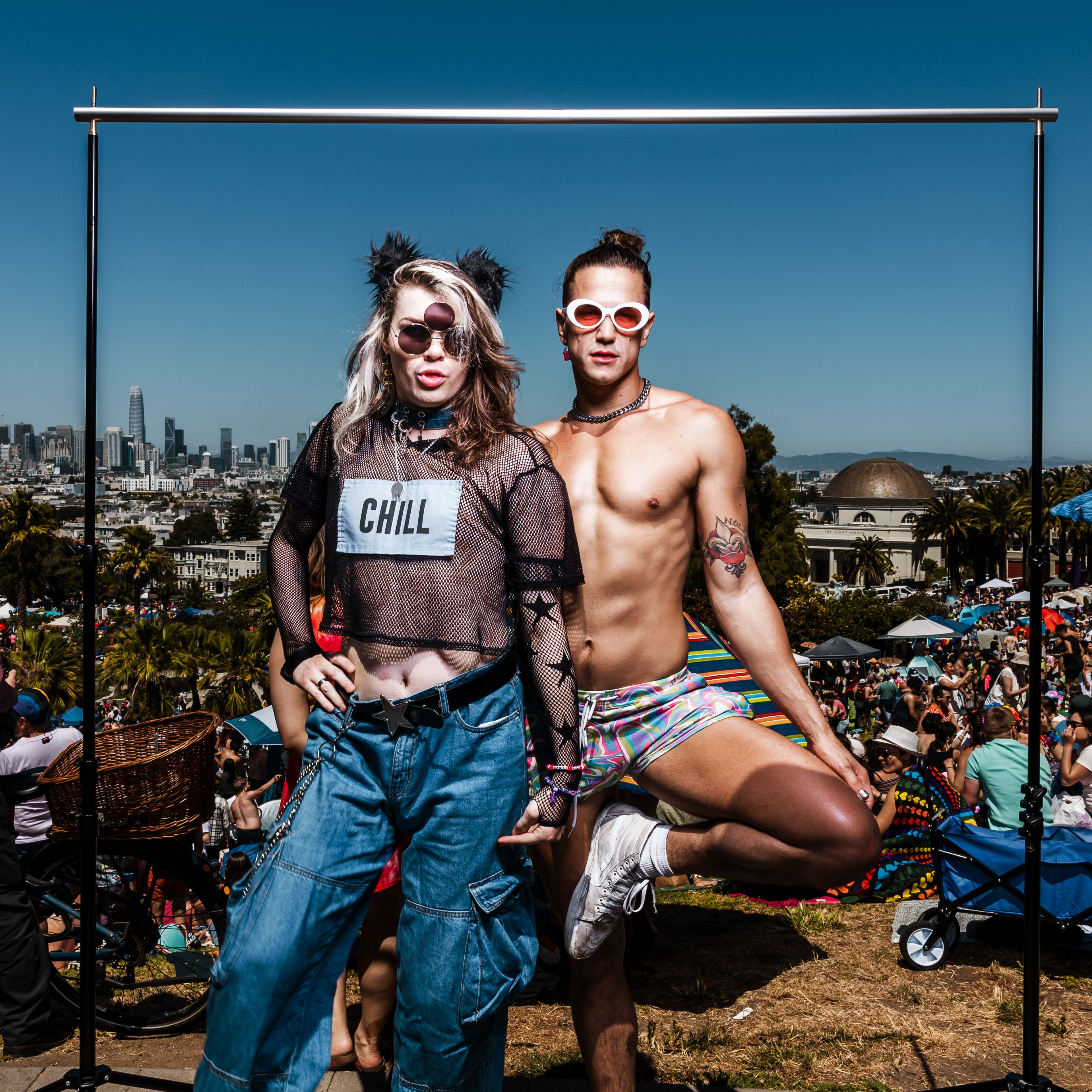 Two individuals pose in colorful outfits at an outdoor event with a city skyline in the background. One wears a &quot;CHILL&quot; shirt, jeans, and cat ears; the other is shirtless in shorts.