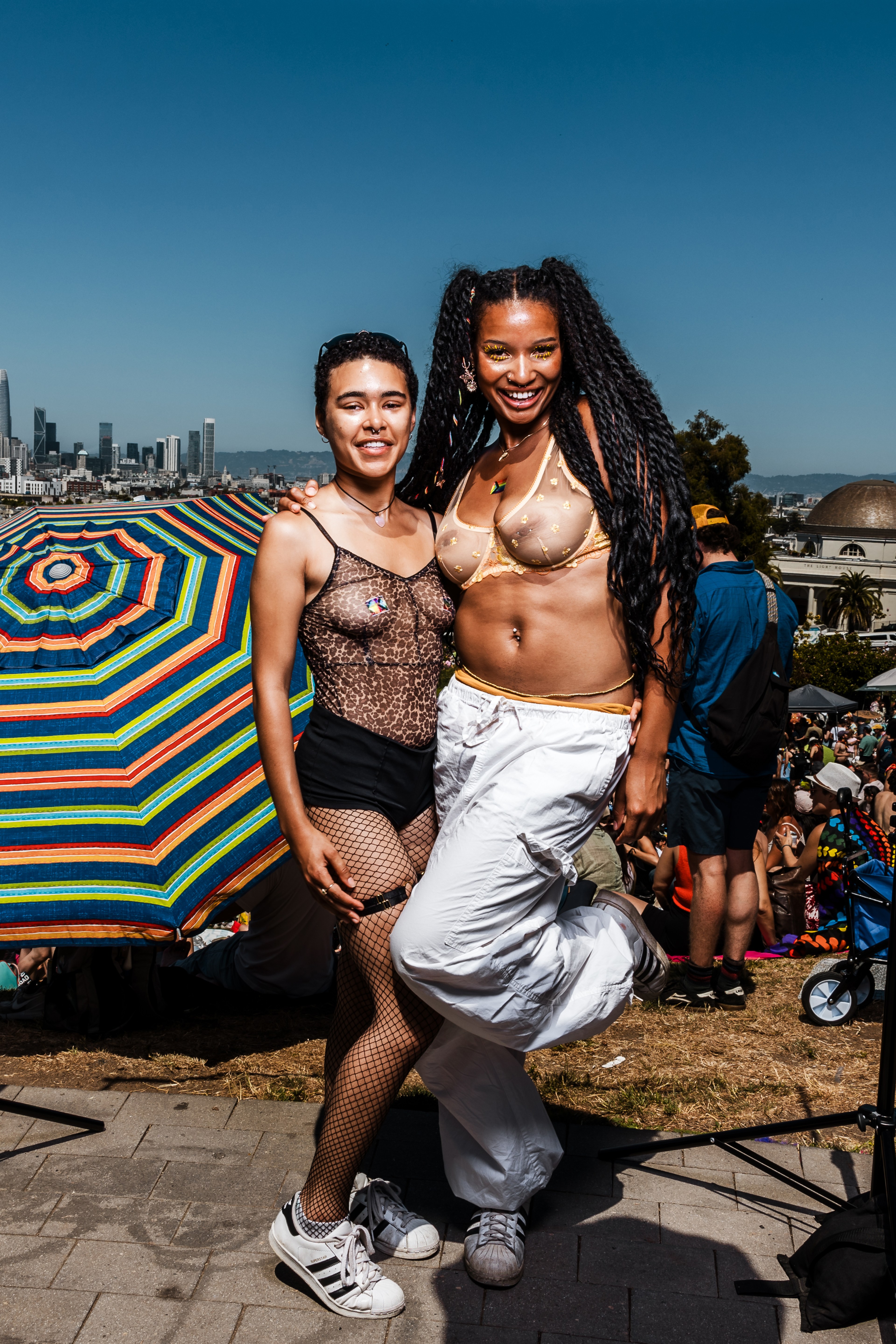 Two people pose cheerfully, one in a sheer leopard print top and fishnet stockings, the other in a sheer gold top and white pants. A colorful umbrella and a cityscape are visible behind them.
