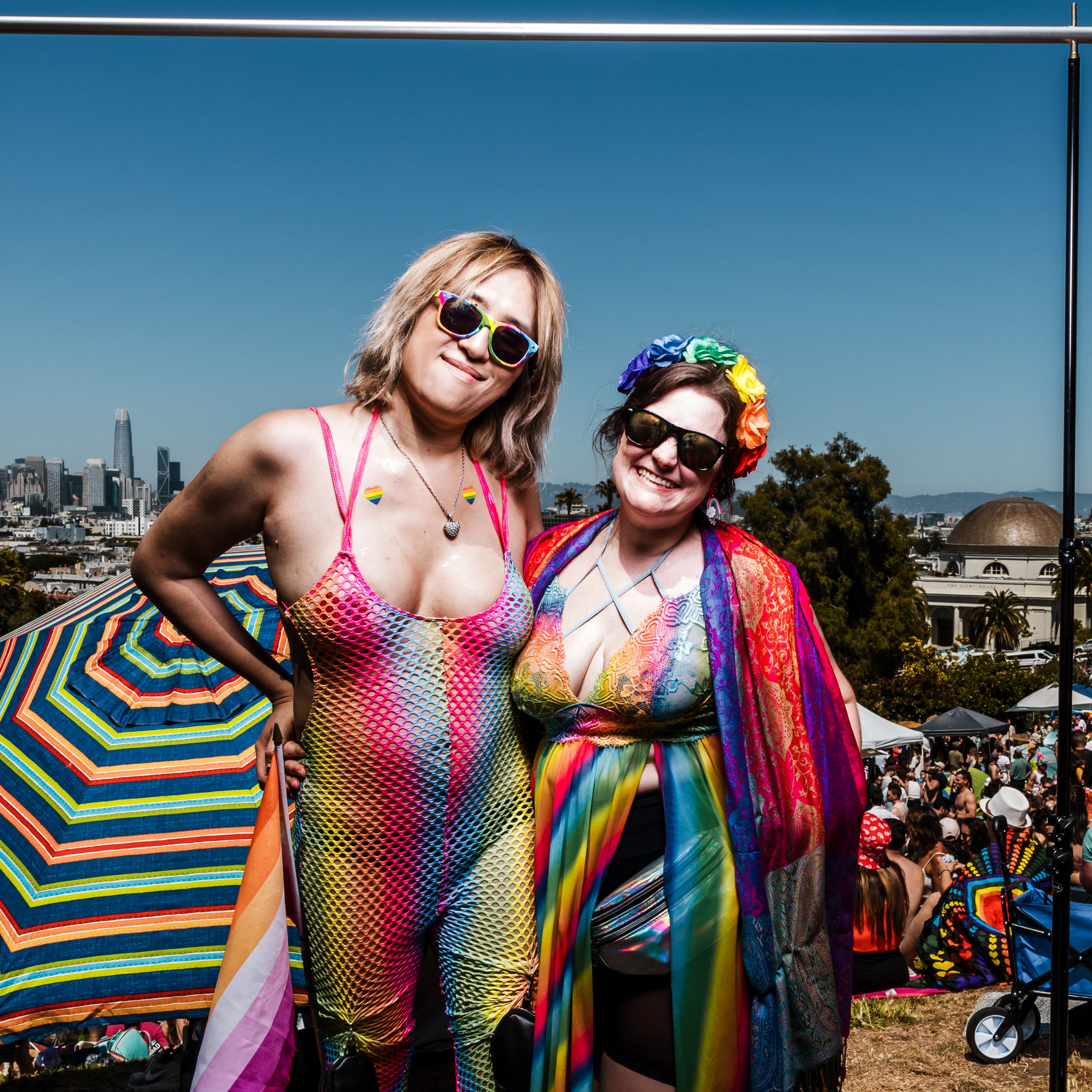 Two people in rainbow clothing and sunglasses stand smiling at a crowded outdoor event with a vibrant cityscape and colorful umbrella in the background.