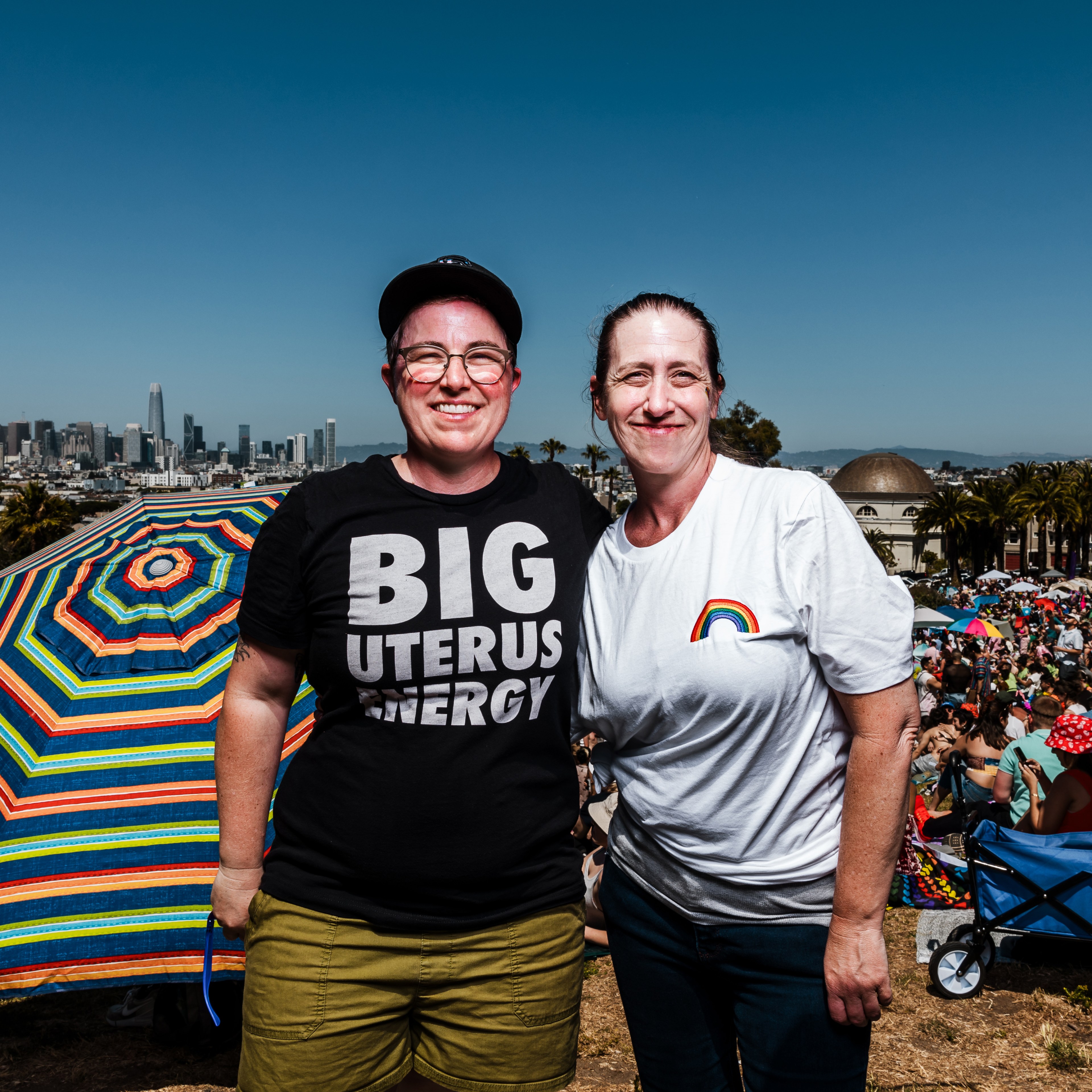 Two people stand smiling in an outdoor setting with a city skyline in the background. One wears a “BIG UTERUS ENERGY” shirt, the other a rainbow t-shirt. A vibrant umbrella and crowd are behind them.