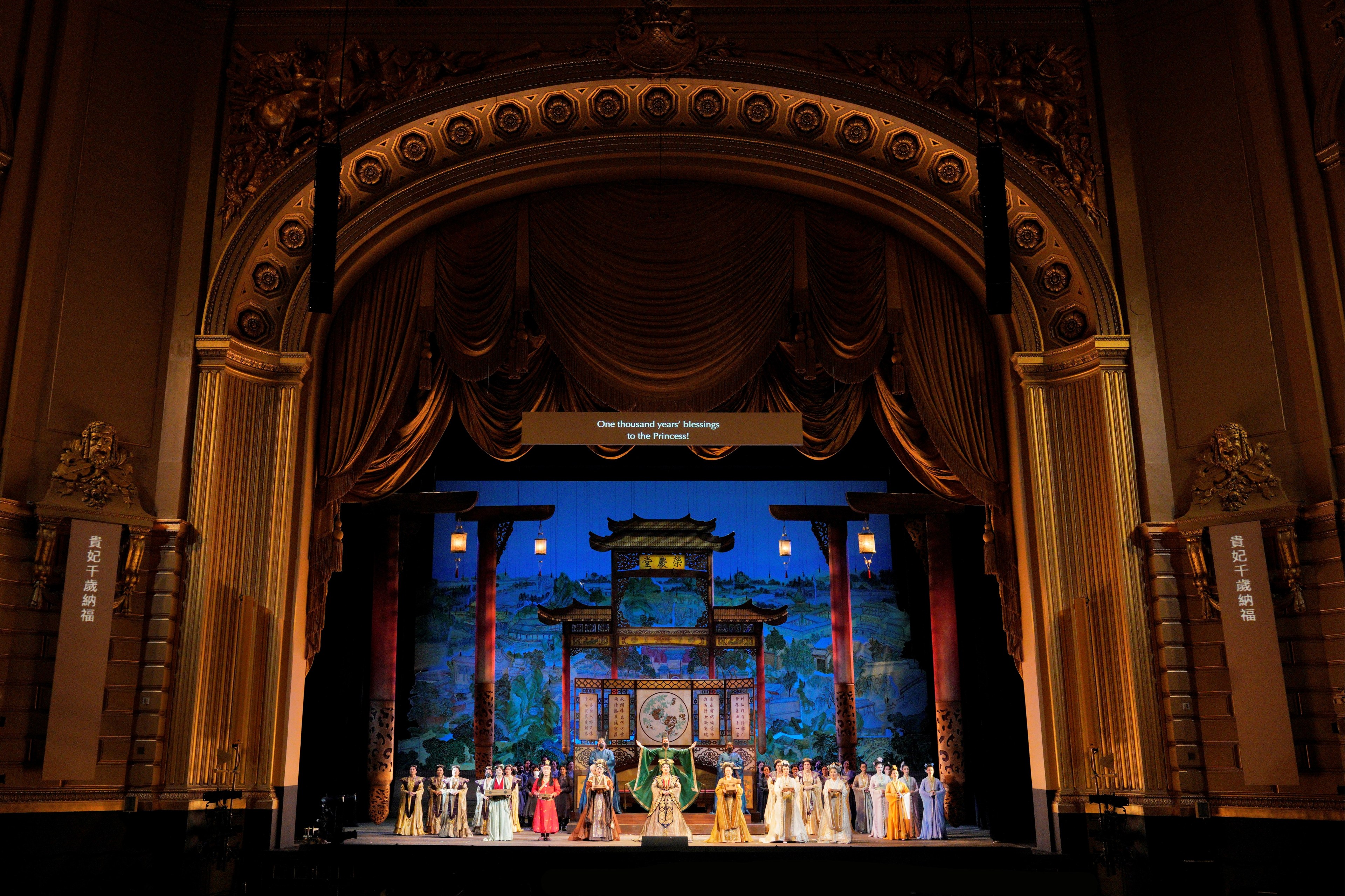 A stage performance with elaborate costumes set in a grand theater. The backdrop features a traditional Asian scene, and the theater has an opulent arch and drapes.