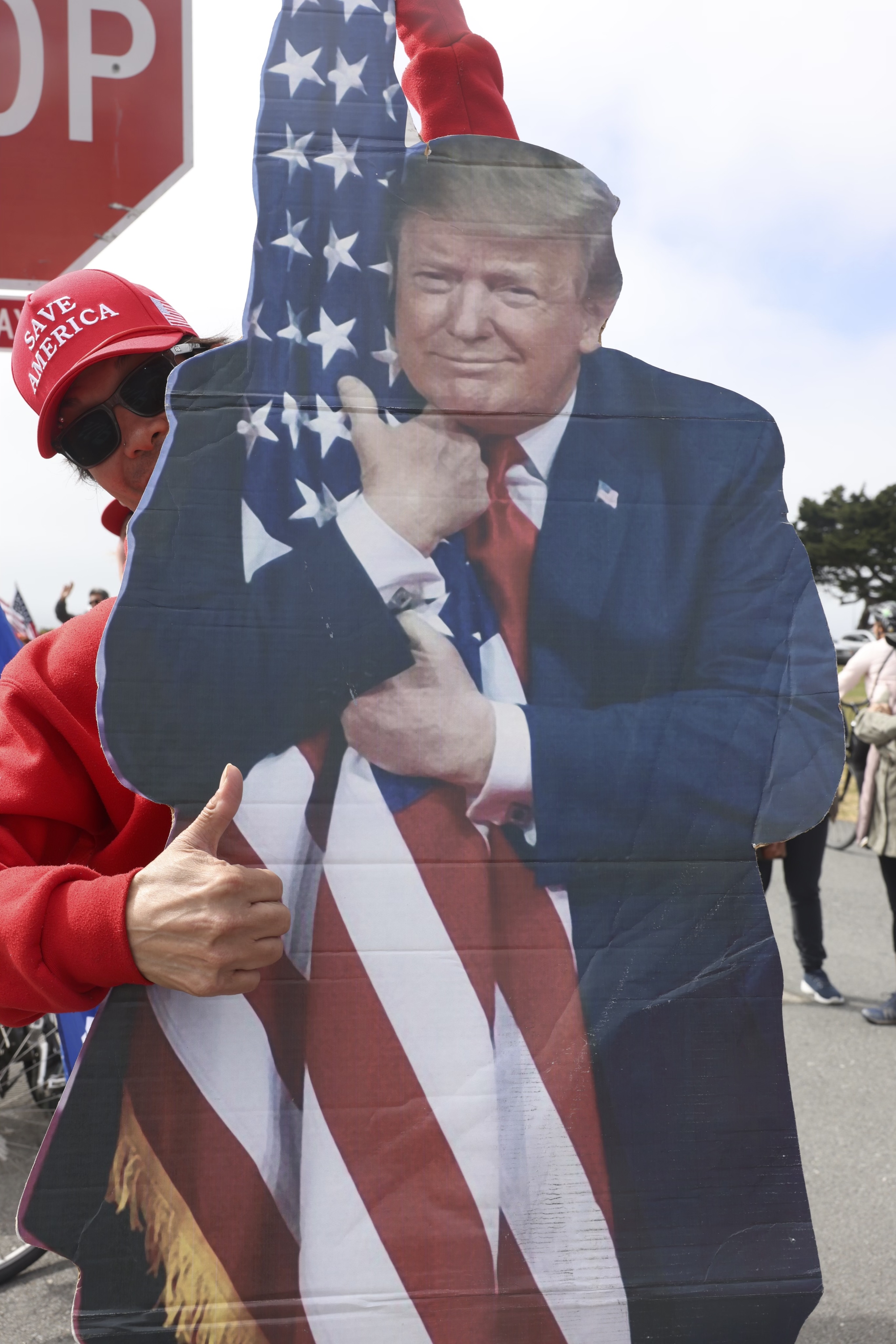 A person in a red &quot;Save America&quot; hat and sunglasses is holding a large cutout of a man hugging an American flag. They show a thumbs-up sign next to a stop sign.