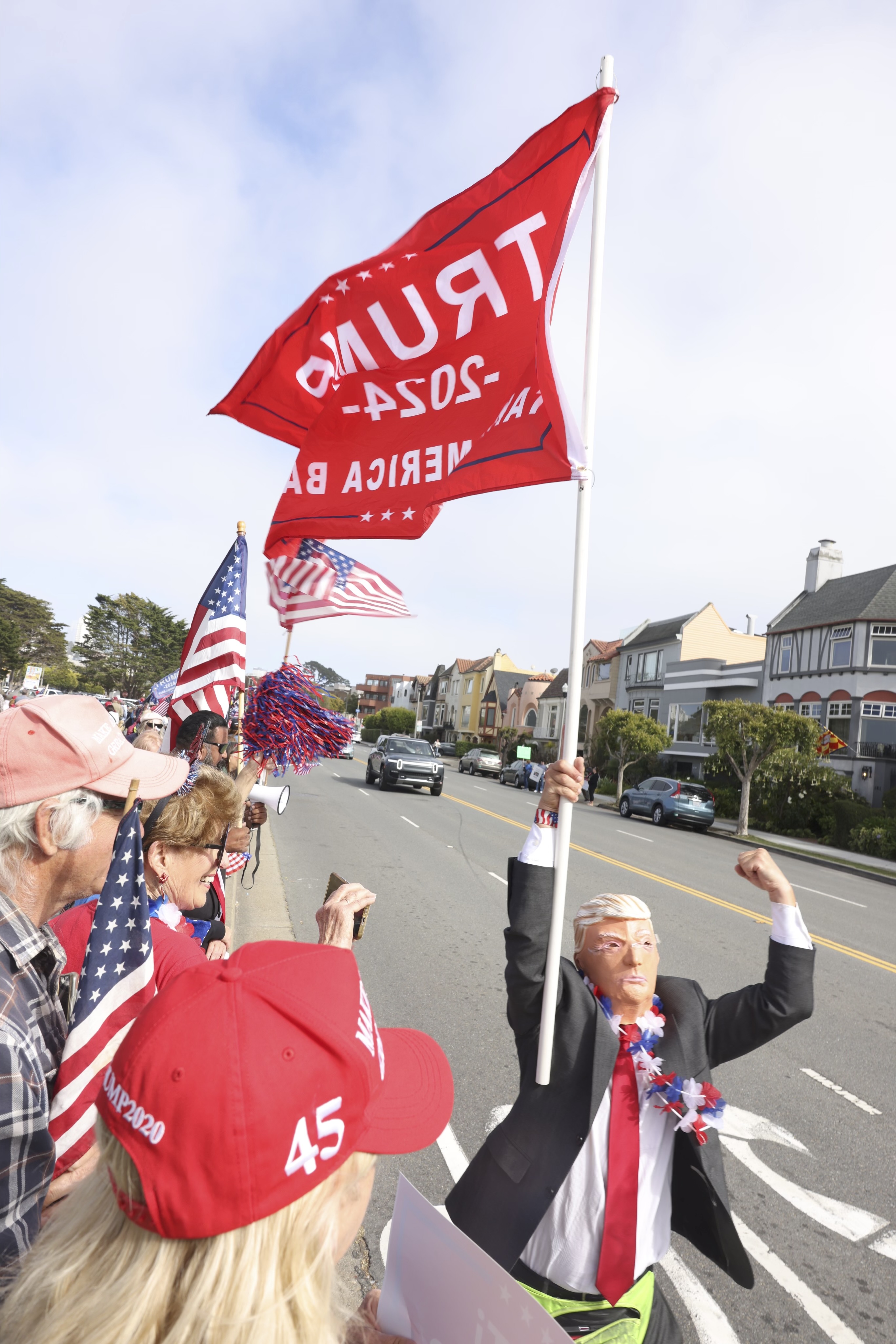 People gather roadside, holding American flags and signs supporting Trump 2024. A person in a Trump mask and suit raises a large red flag with &quot;Trump 2024&quot; printed on it.