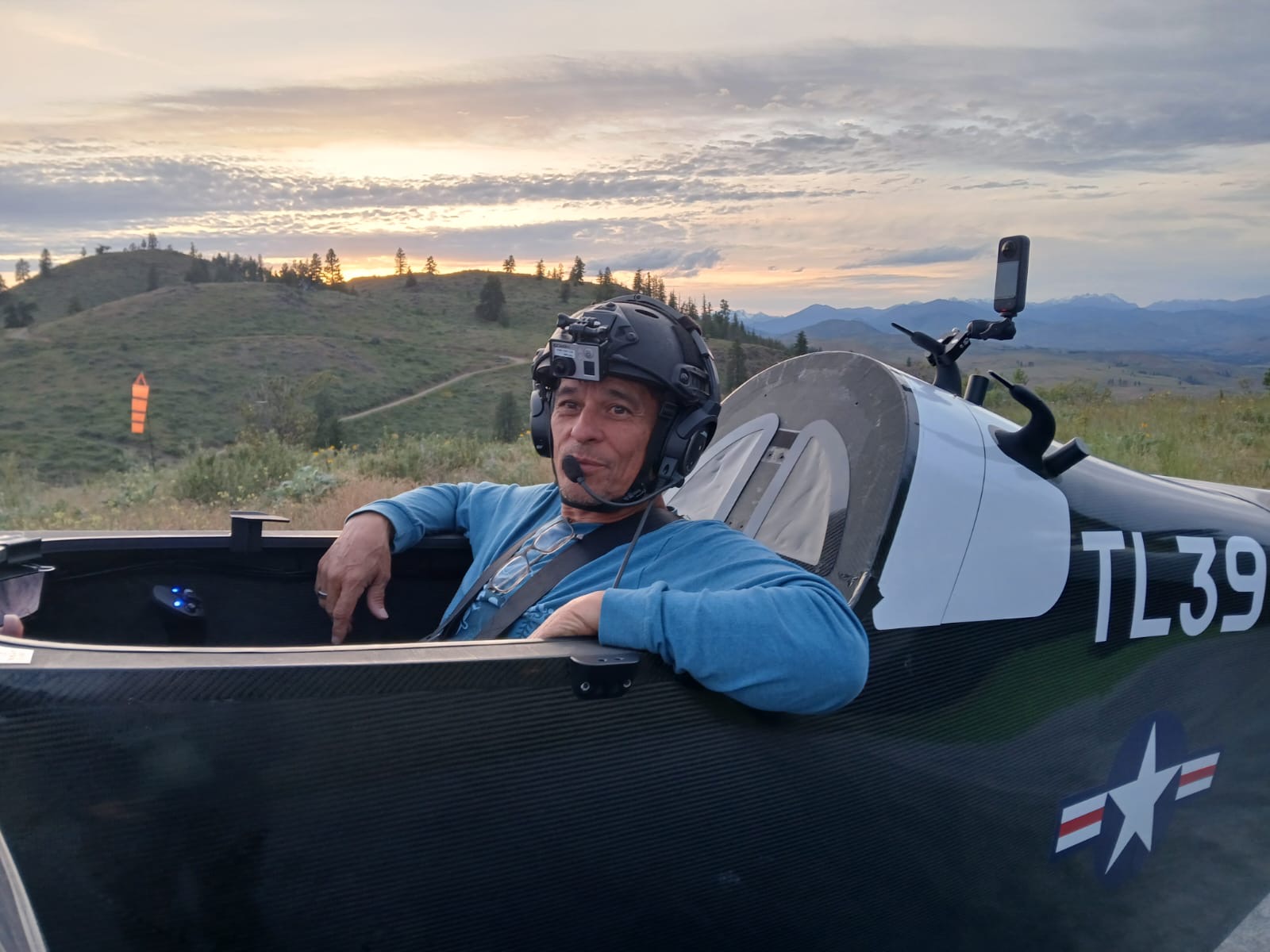 A person wearing a helmet with a mounted camera and headset sits in a black glider cockpit marked with &quot;TL39.&quot; The background shows a hilly, scenic sunset.