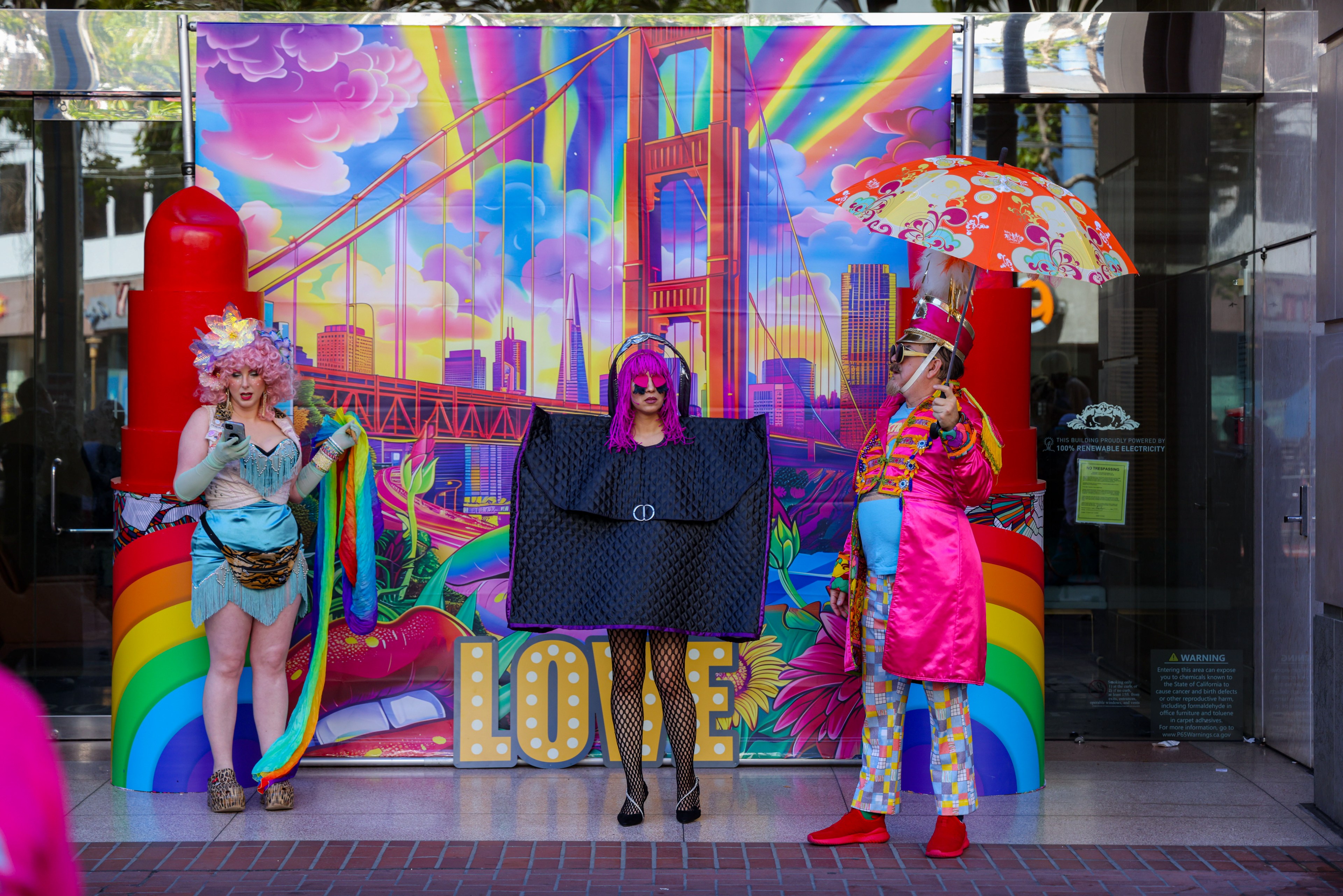 Three people in flamboyant, colorful costumes pose in front of a vibrant mural of the Golden Gate Bridge. The mural features rainbows, clouds, and the word &quot;LOVE.&quot;