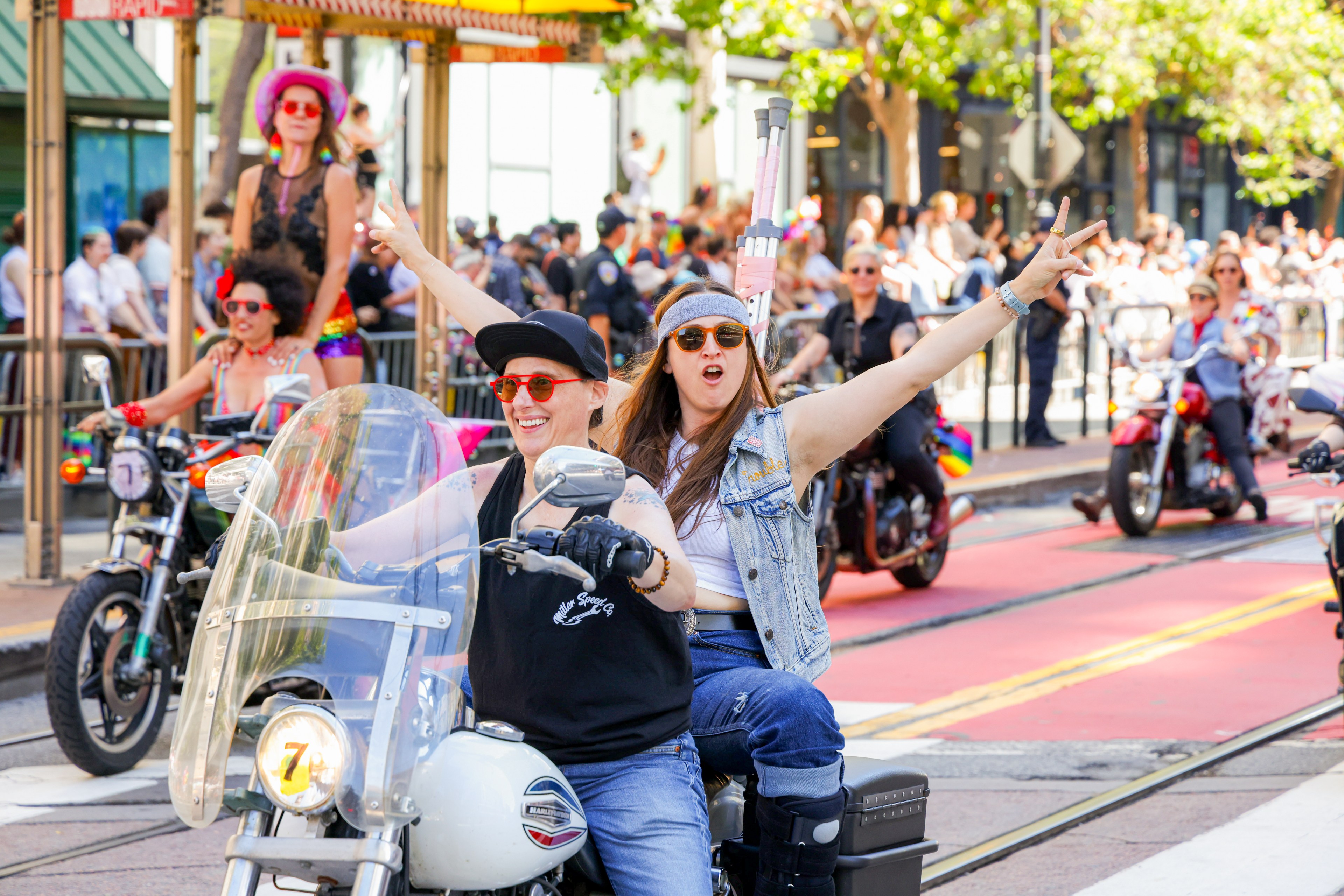 A lively parade features jubilant individuals on motorcycles. Two women on one motorcycle lead, arms raised in celebration, with colorful attire and a cheerful crowd in the background.