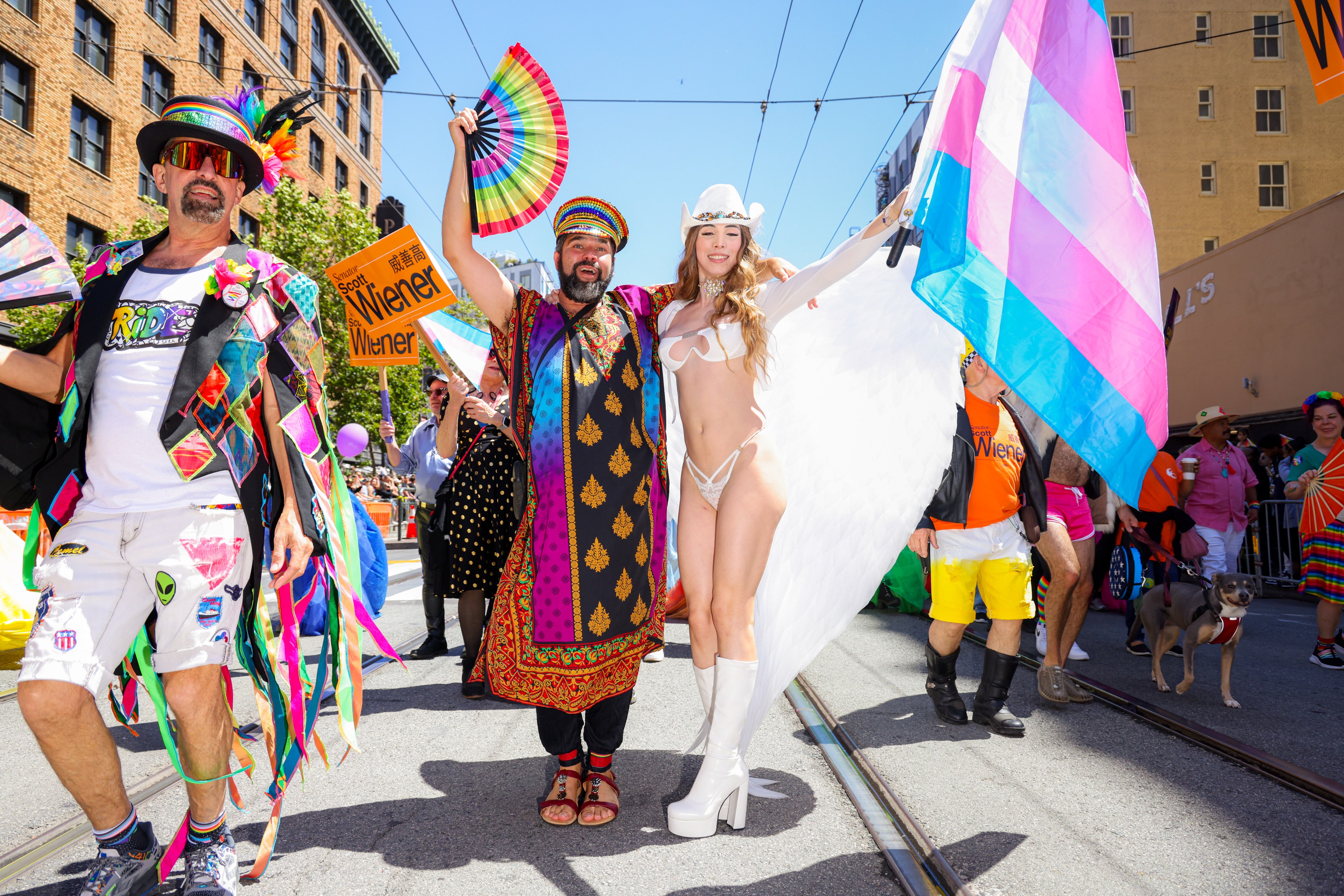 A colorful parade with people in vibrant costumes; one person wears black and white with rainbow fans, another wears a patterned robe, and a third in white lingerie has angel wings.