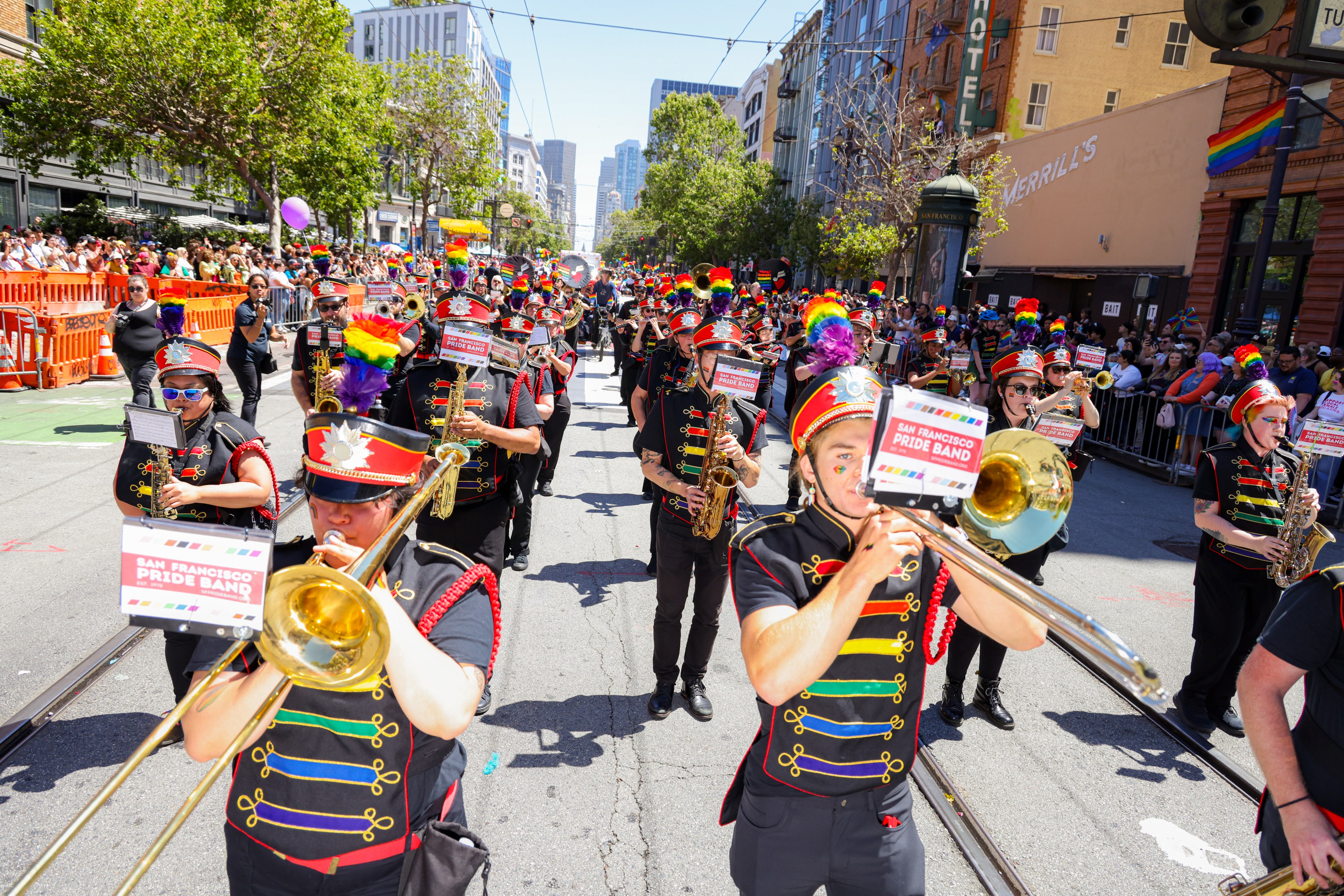 A brass band dressed in black and red uniforms with rainbow accents marches down a city street adorned with Pride flags, while crowds cheer from behind barricades on a sunny day.
