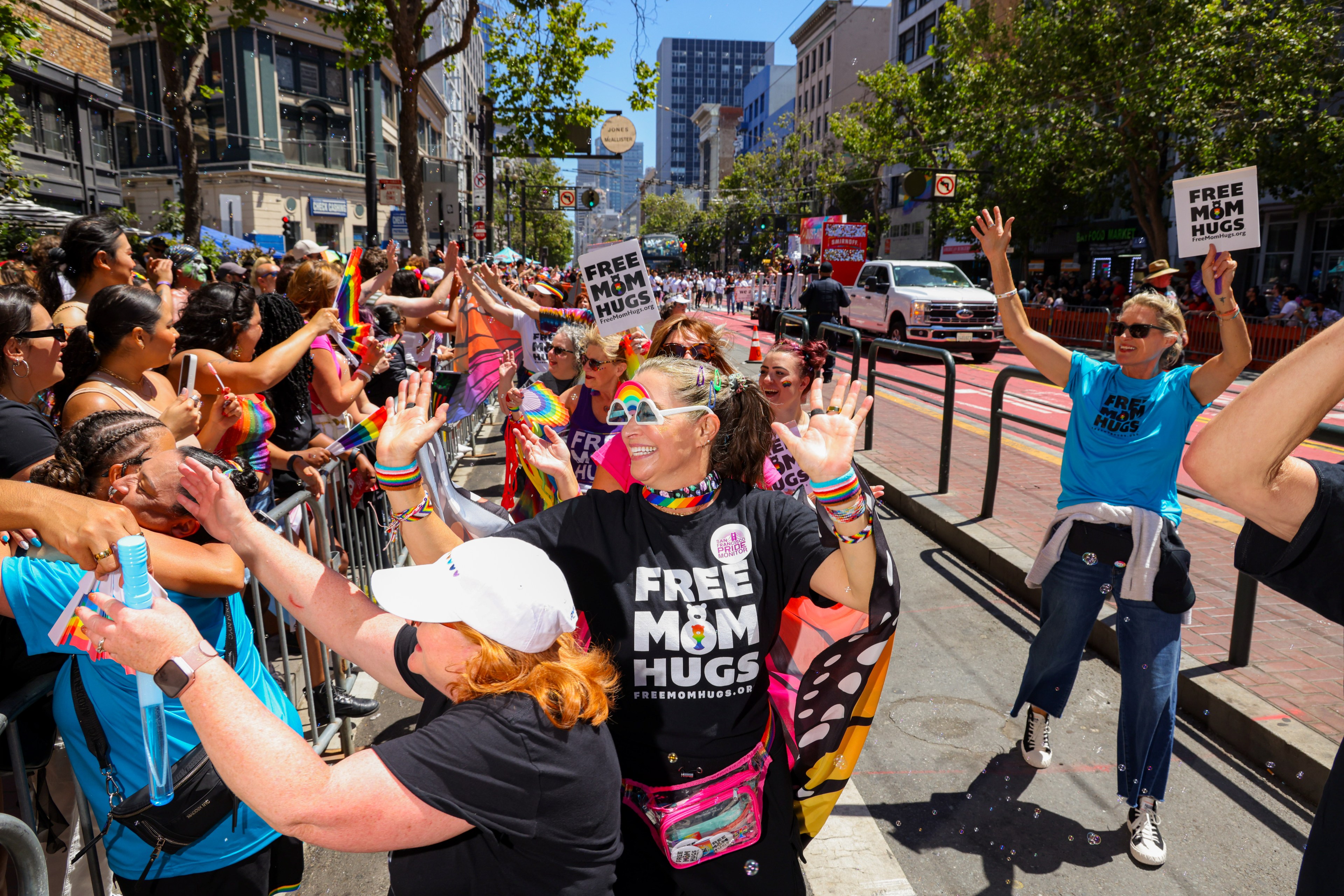 A group of people in colorful attire and &quot;Free Mom Hugs&quot; shirts interact joyfully with a cheering crowd behind a barrier on a sunlit urban street.