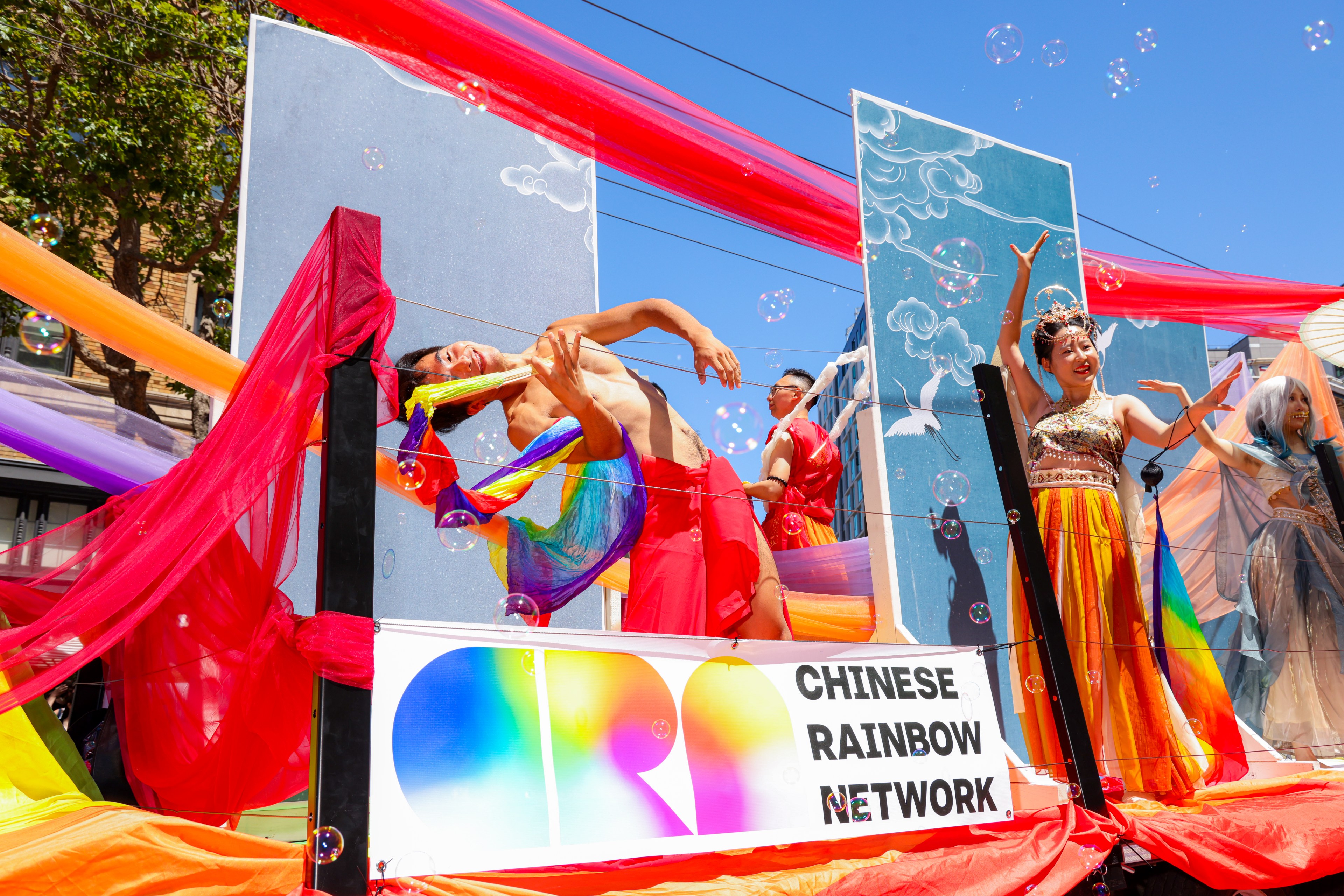 A colorful parade float features dancers in vibrant costumes and rainbow fabric under a clear sky. The sign reads &quot;CHINESE RAINBOW NETWORK&quot; amid floating bubbles.