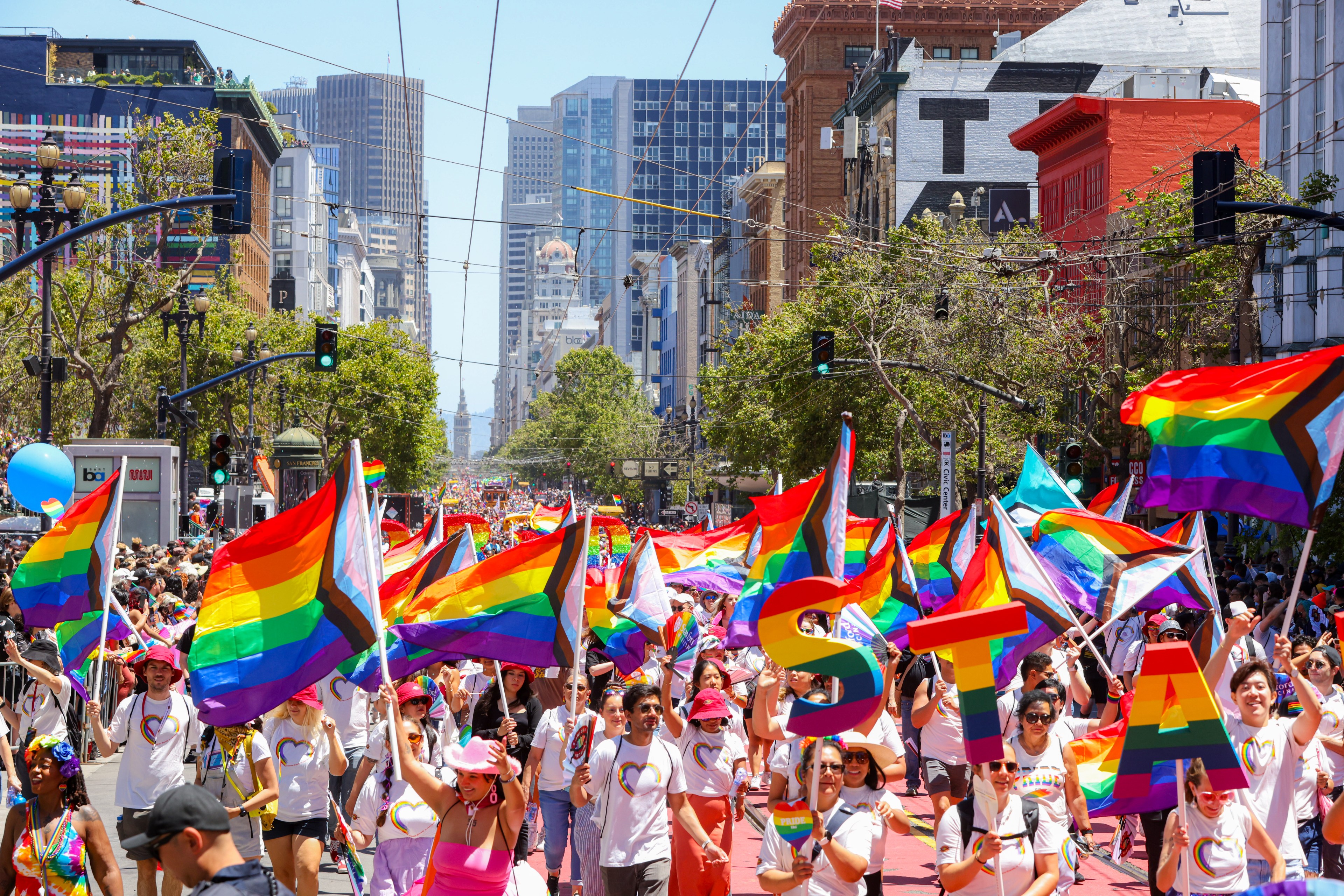 A vibrant parade fills a city street with people waving rainbow flags and wearing colorful outfits. Tall buildings and greenery are in the background.