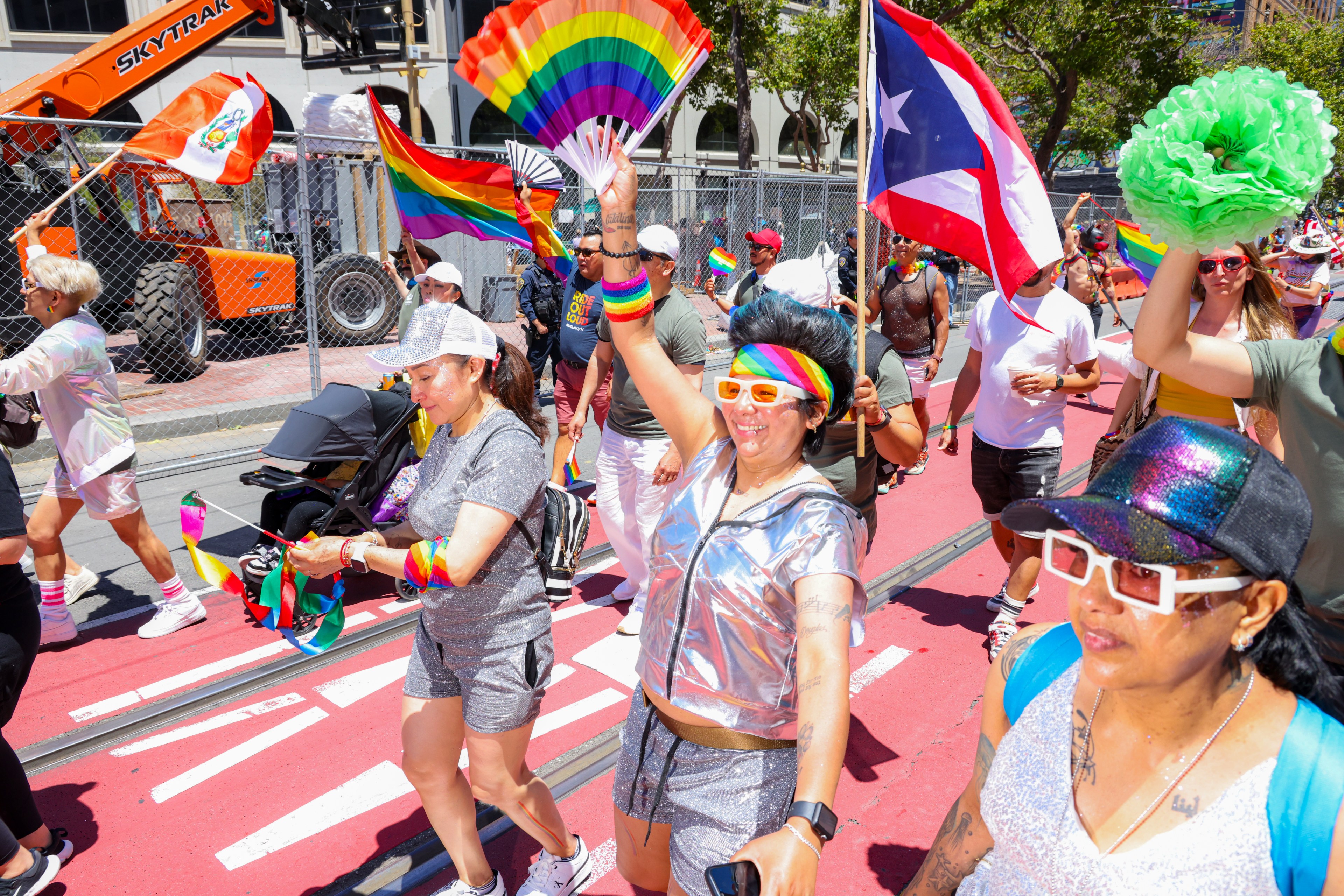 People in colorful outfits are joyfully participating in a Pride parade, waving rainbow flags and banners, on a brightly sunlit street.