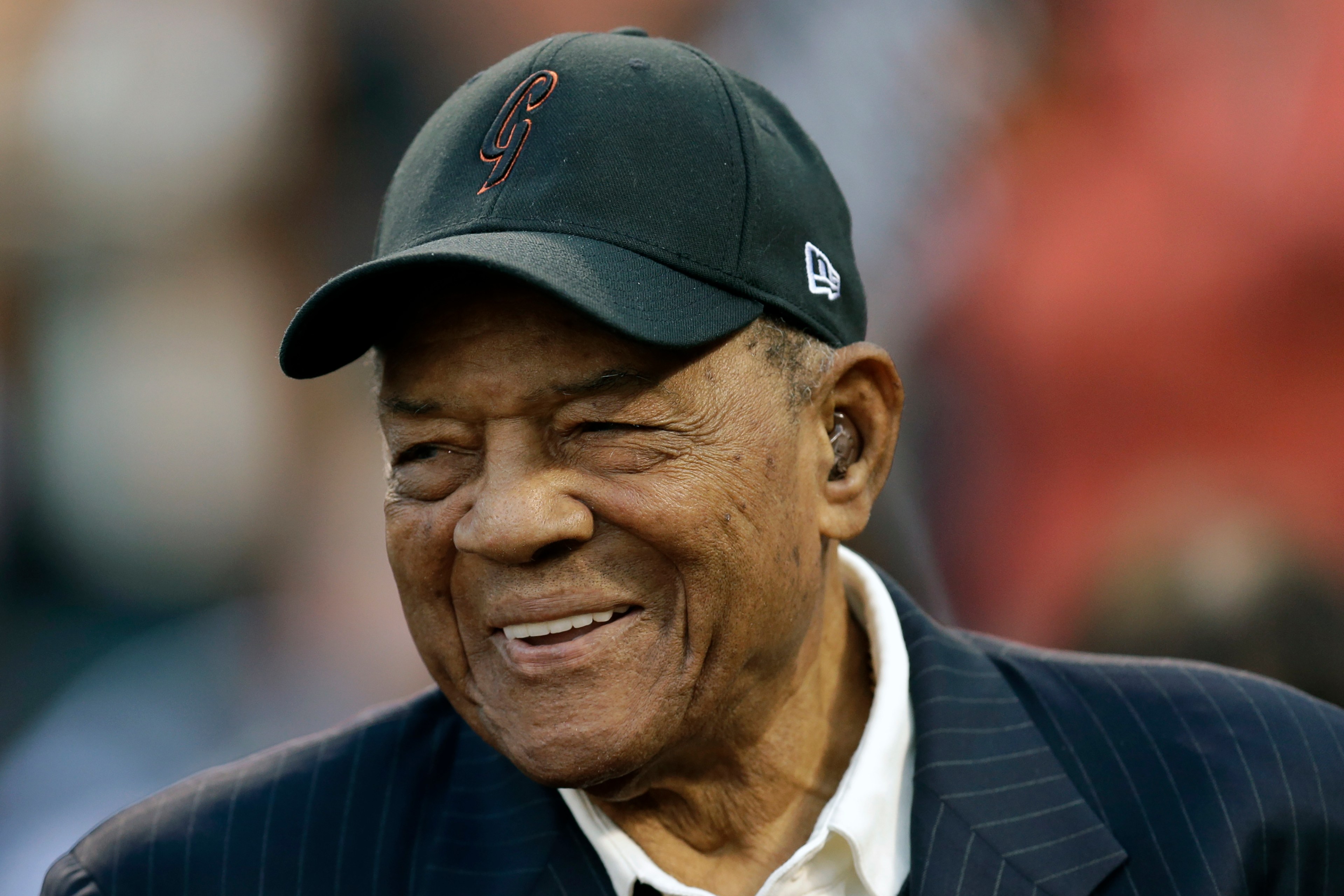 An elderly man with a warm smile is wearing a black baseball cap and a dark striped suit jacket over a white shirt. The background is blurred.