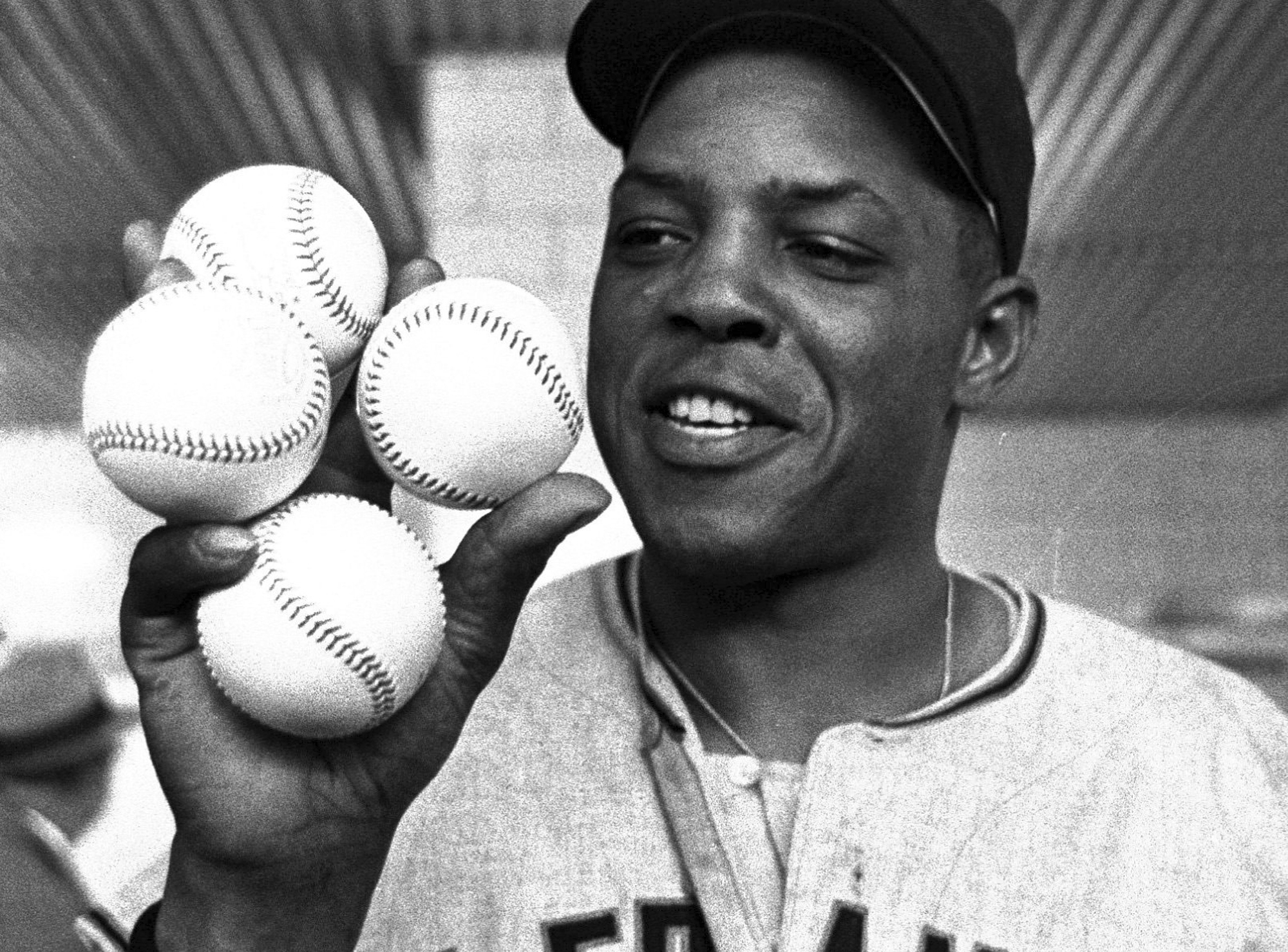 A baseball player holds five baseballs in one hand, smiling. He wears a baseball cap and jersey with visible letters, and the background is an indoor setting.