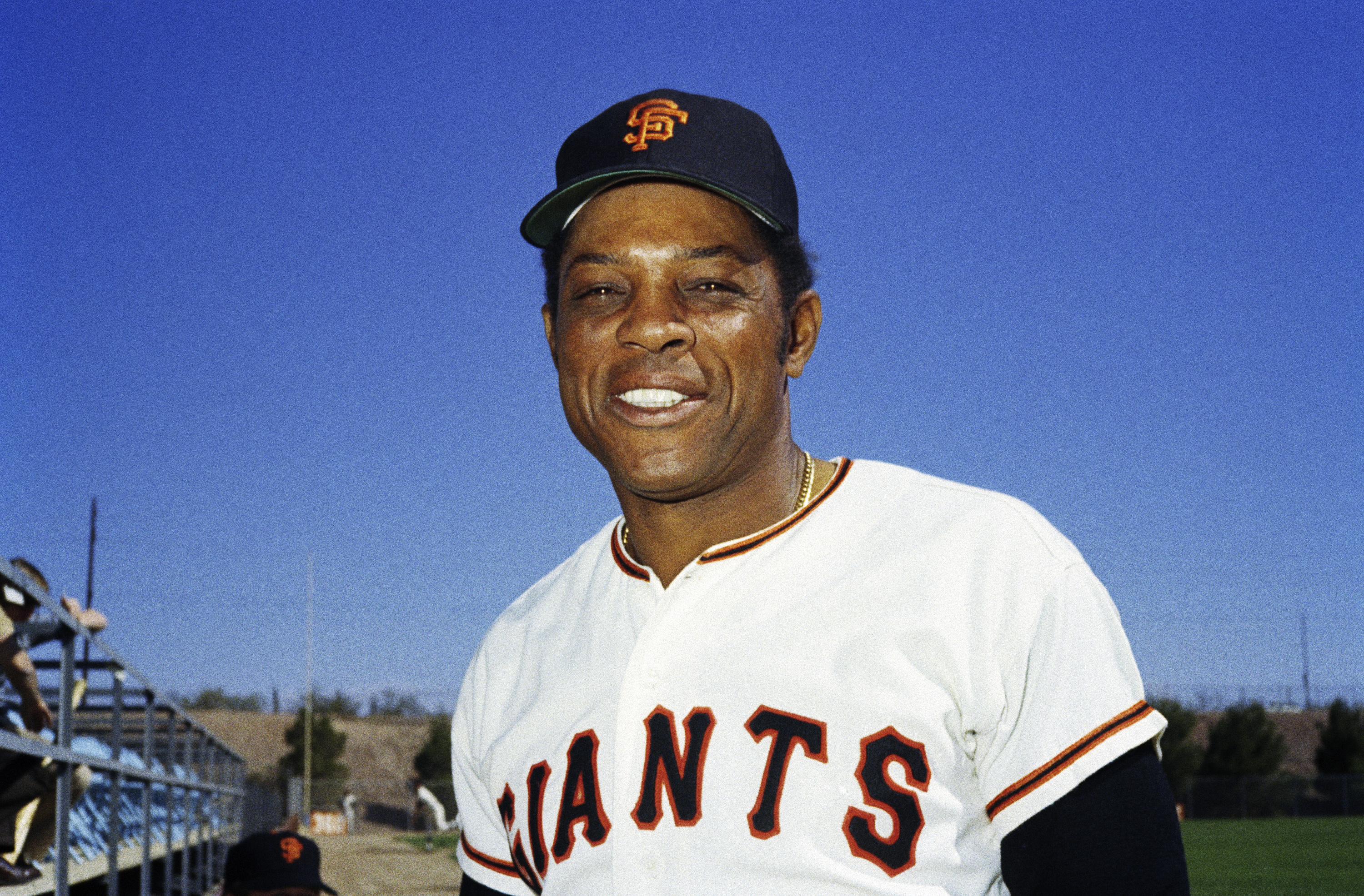 A smiling man wearing a San Francisco Giants baseball uniform stands outdoors against a clear blue sky, with bleachers and a baseball field in the background.