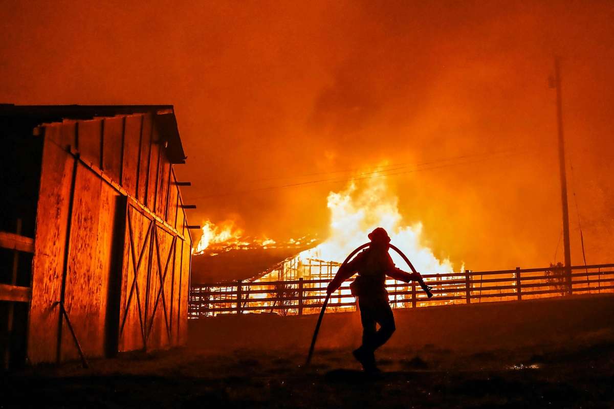 A firefighter carries a hose in front of a large barn engulfed in intense flames, with a smoke-filled, orange sky above.