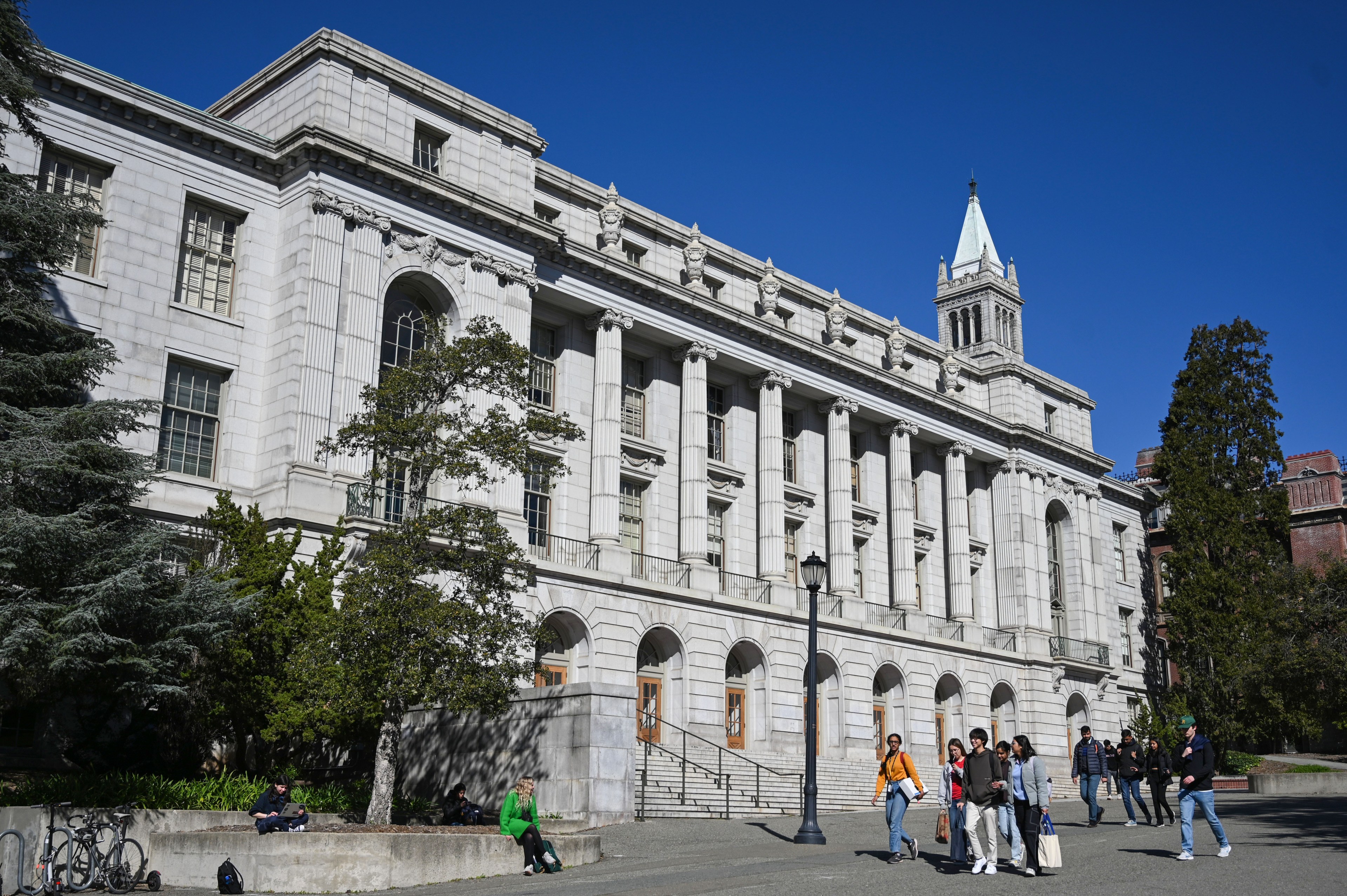 A group of people walk and sit near a large, white stone building with columns, arched doorways, tall windows, and a clock tower under a clear blue sky.