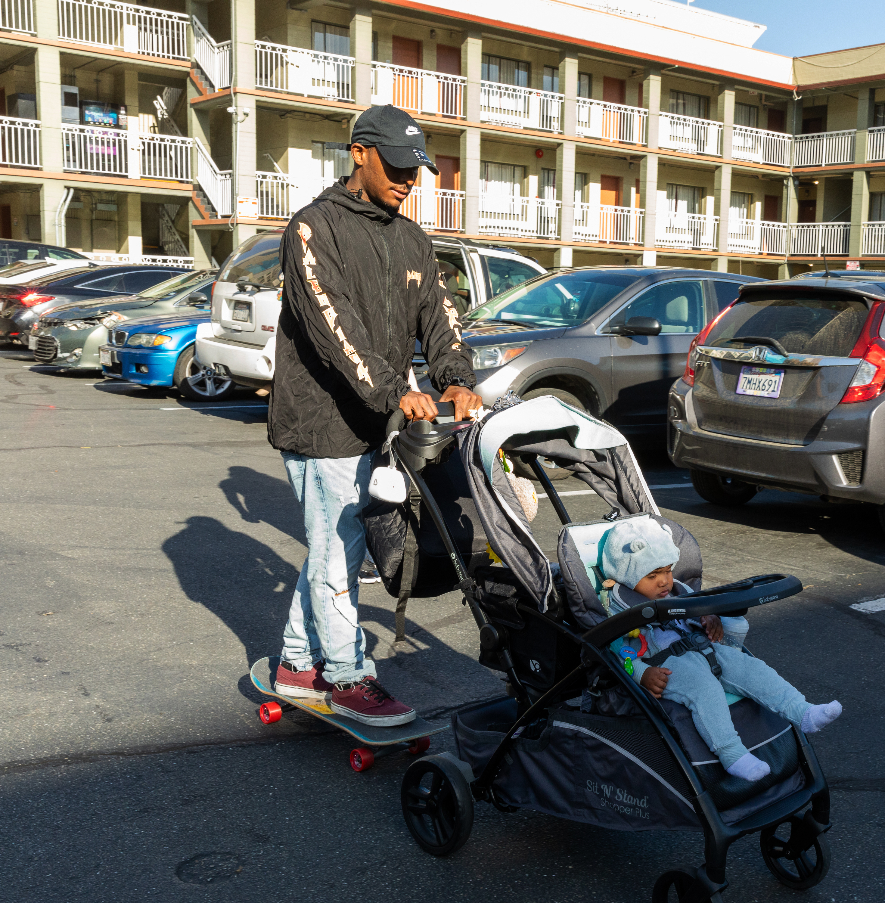 A man in a black jacket rides a skateboard while pushing a baby in a stroller through a car park, with a multi-story building in the background.