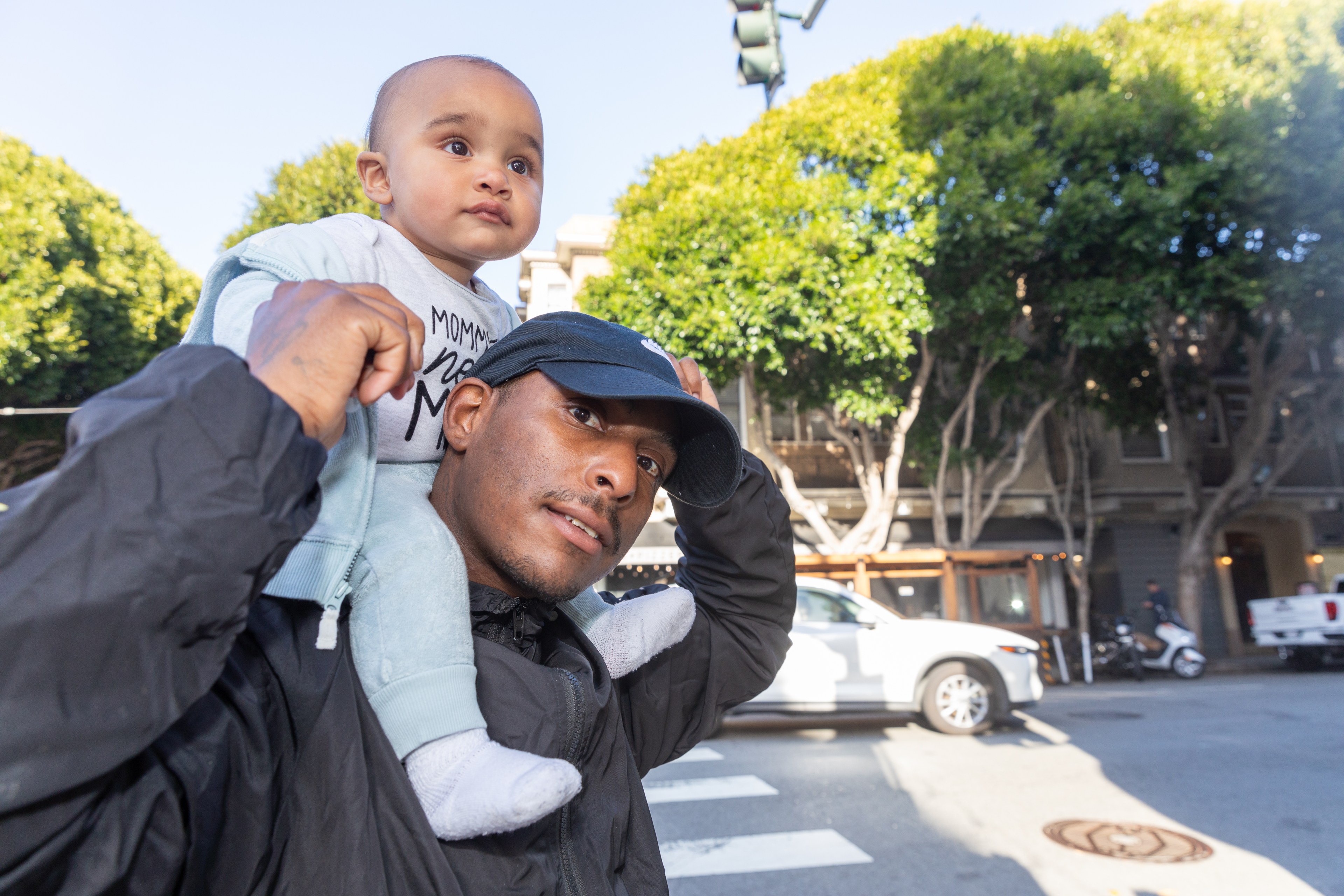 A man is carrying a baby on his shoulders while outdoors. They stand against a background of trees, vehicles, and a building. The baby wears a shirt that reads "Mommy's New Man."