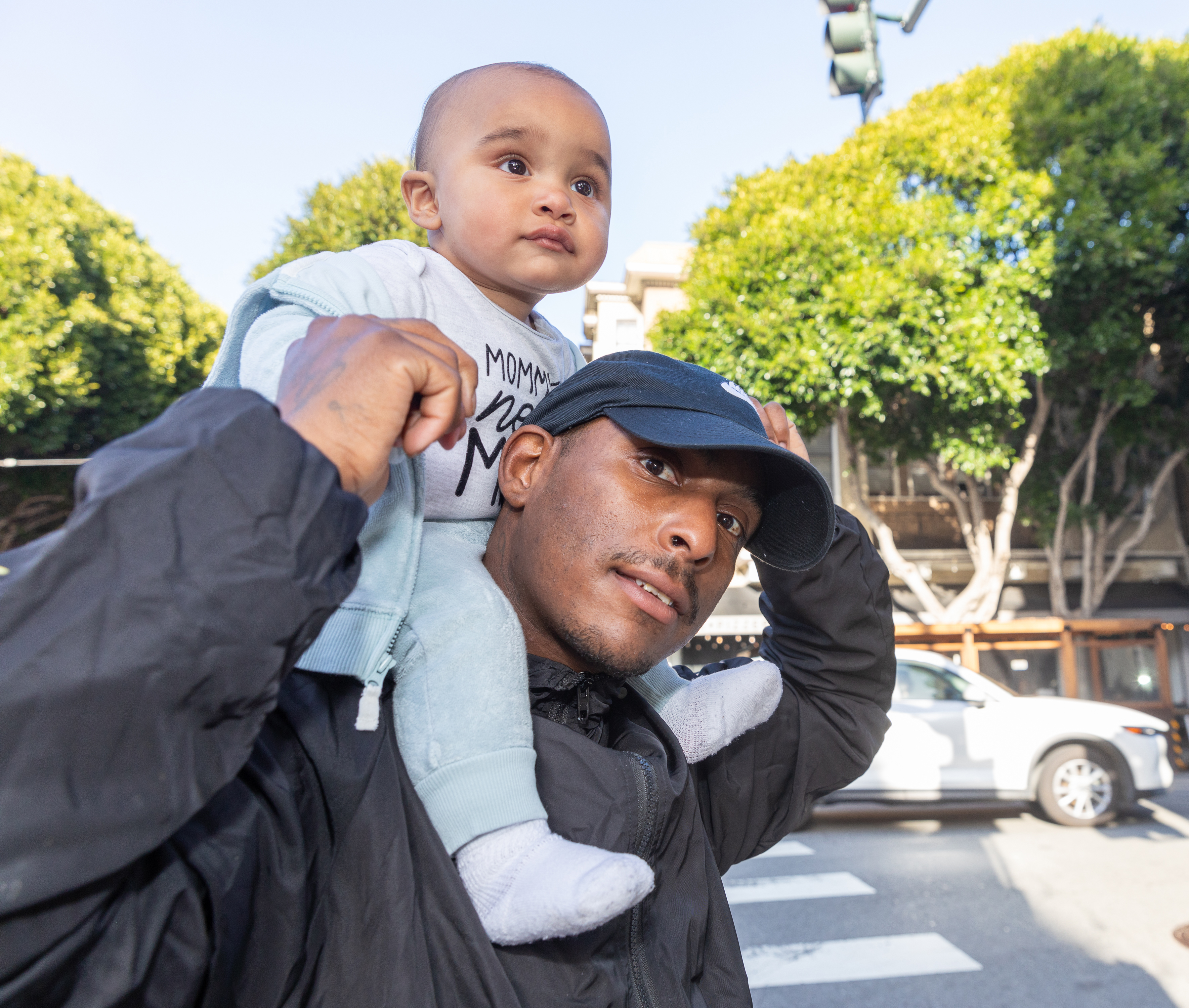 A man is carrying a baby on his shoulders while walking outside. The man wears a black cap and jacket, and the baby has an alert expression, with greenery and cars in the background.