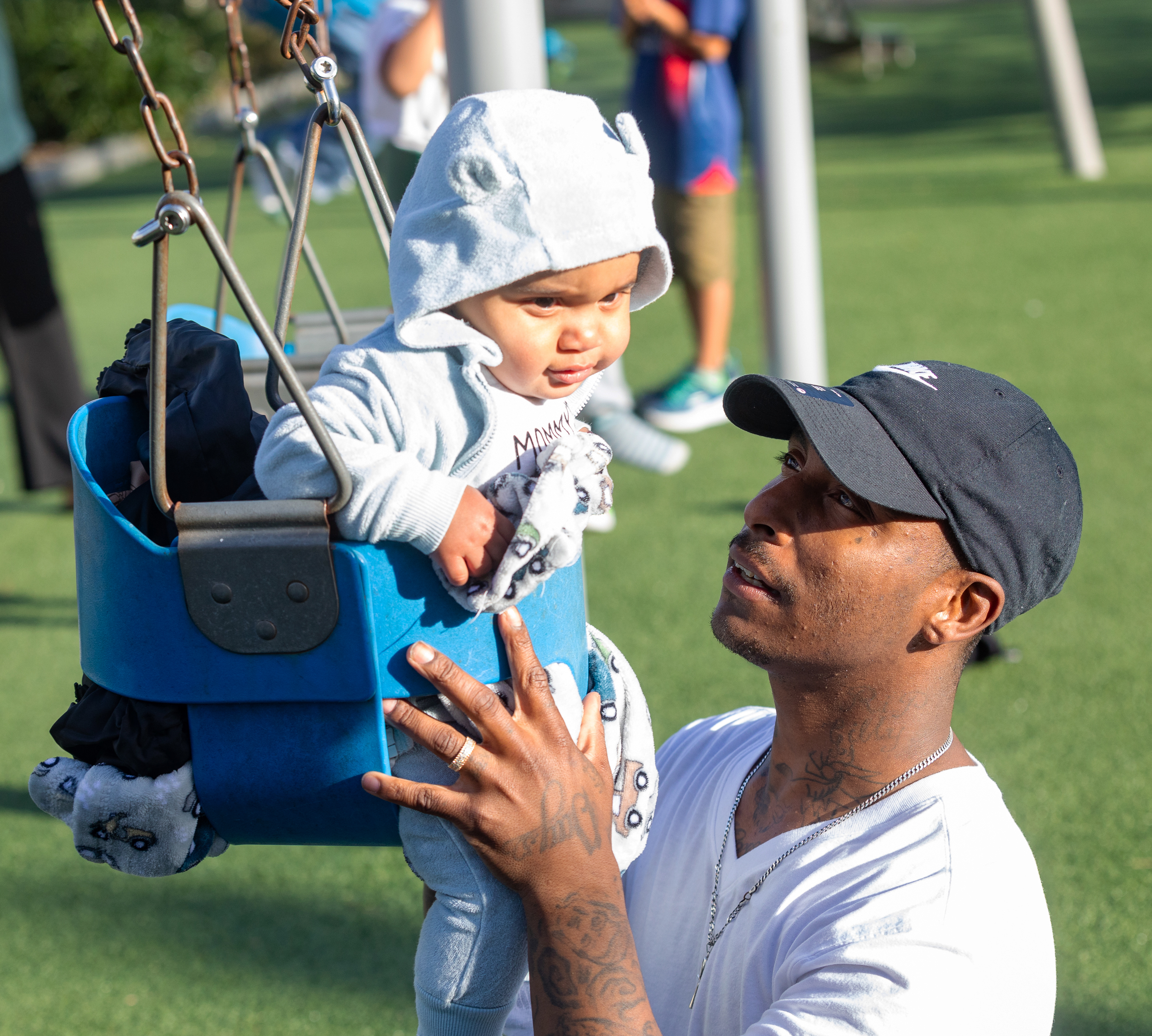 A man pushes a baby in a blue swing. The baby, wearing a hoodie, smiles while the man holds the swing, looking at the baby with care.