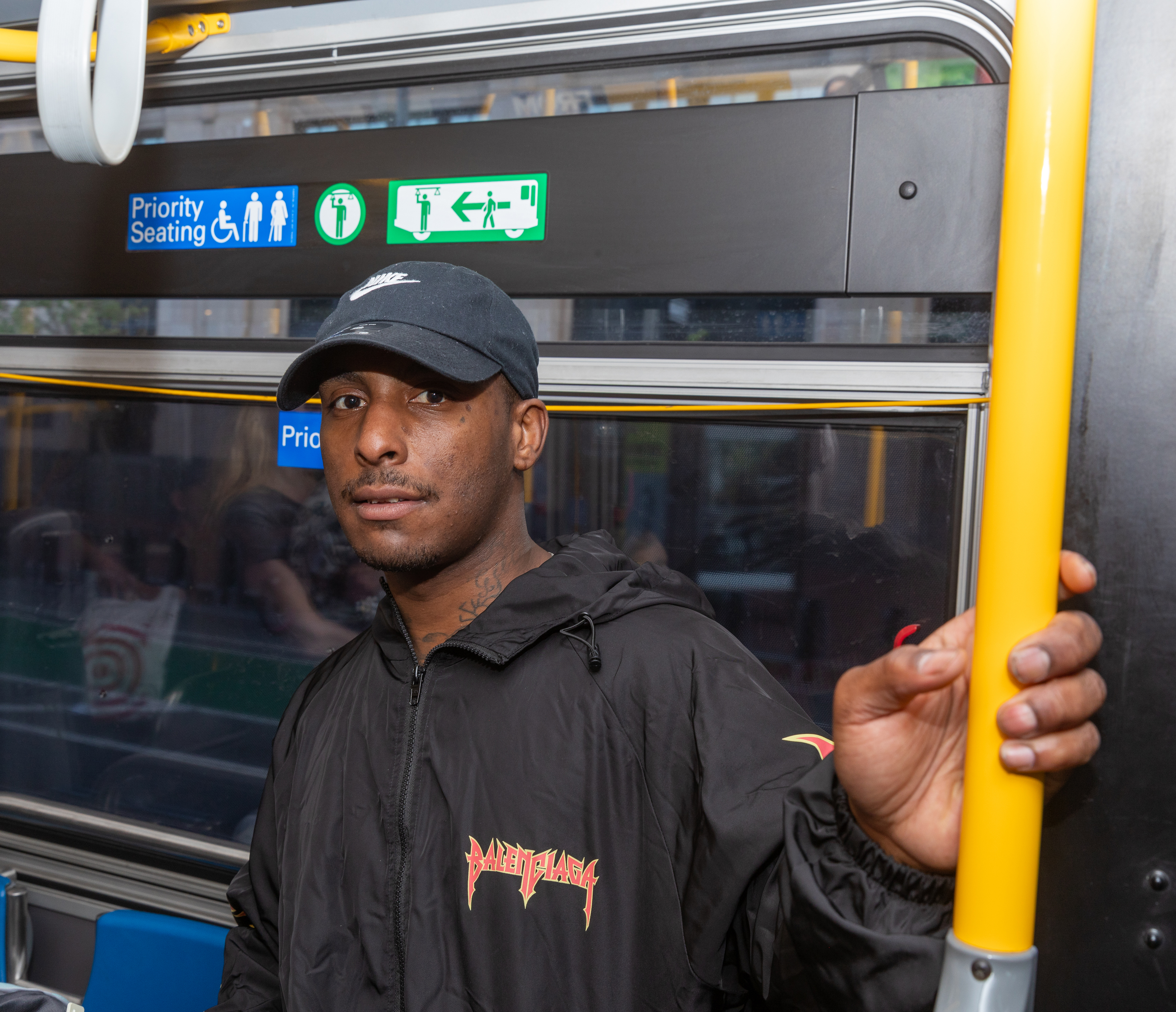 A man wearing a black jacket and cap is standing inside a bus, holding a yellow pole. There are signs above for priority seating and emergency exits.