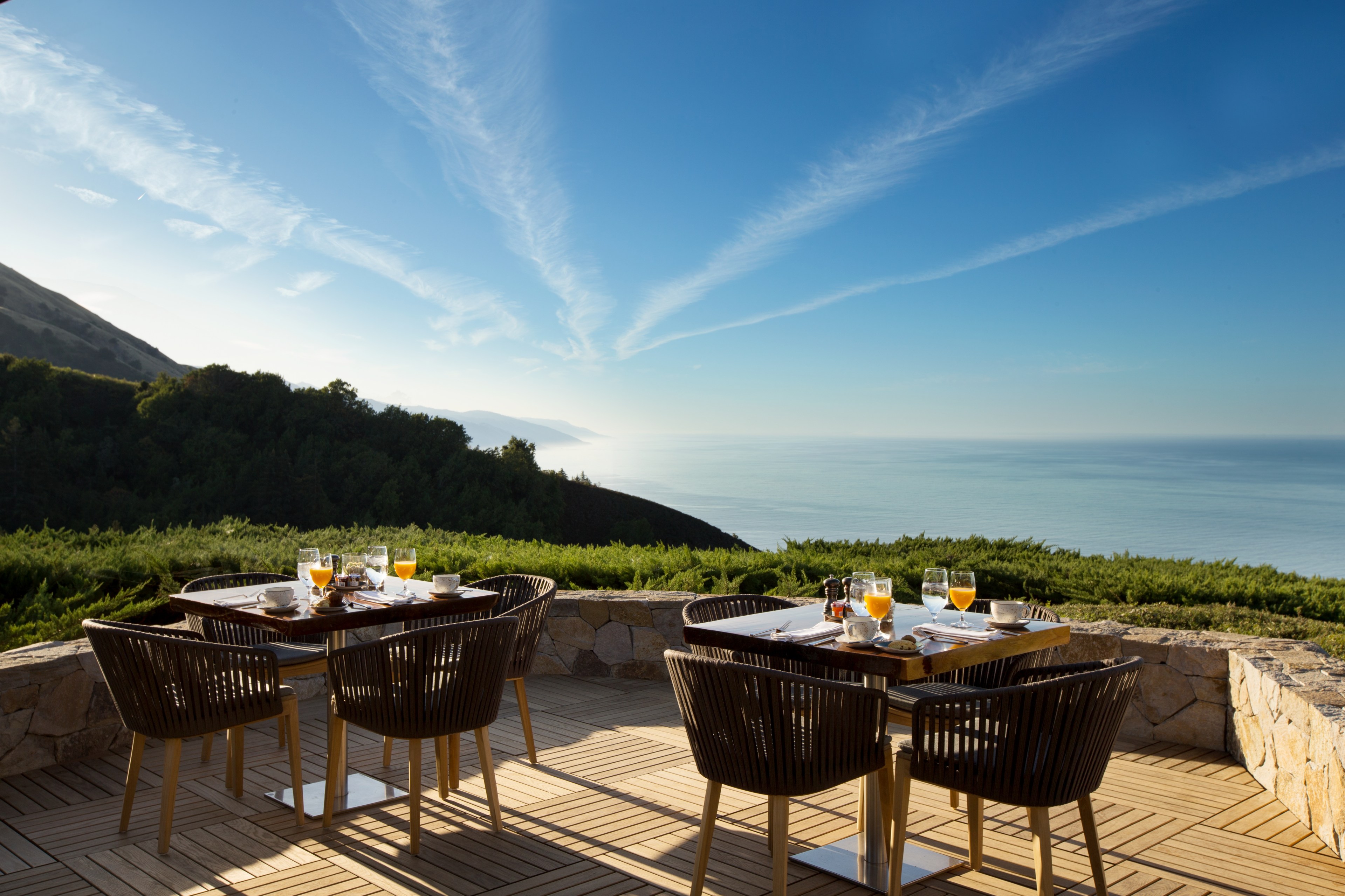 A scenic outdoor dining area overlooks a lush green landscape and a distant sea under a clear blue sky with wispy clouds, with two tables set for breakfast.